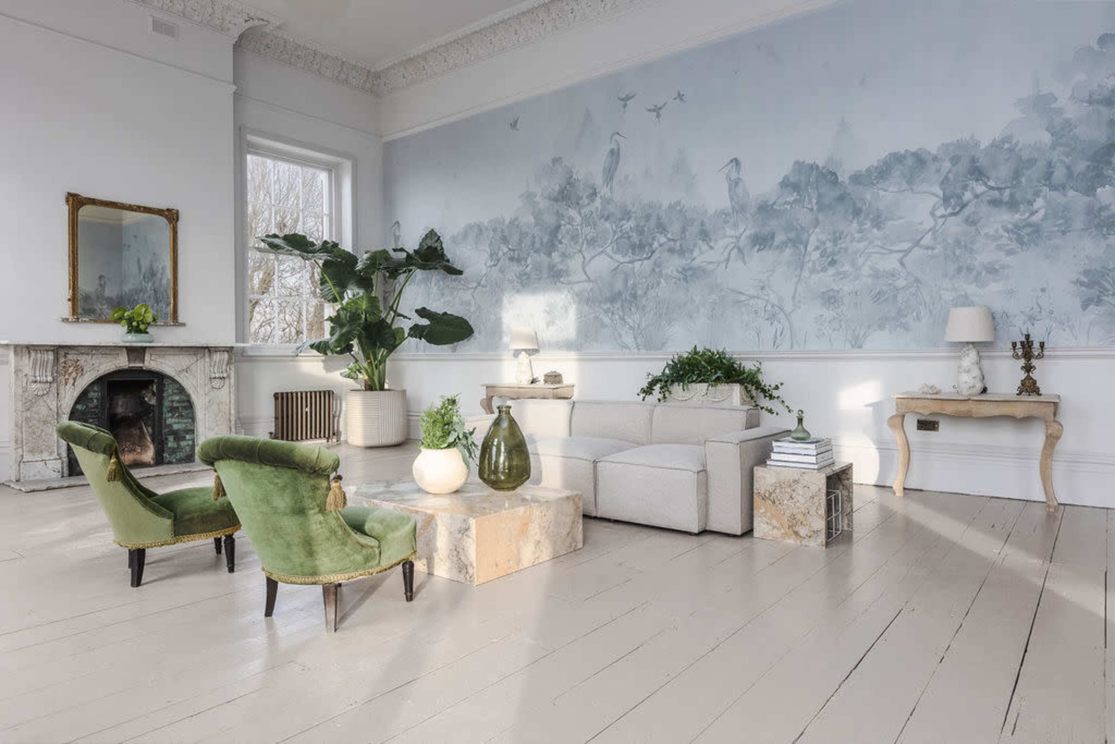 A bright, airy living room featuring a light-colored sofa, two green chairs, a large mural on the wall, and a marble coffee table, with plants and decorative items arranged throughout the space.