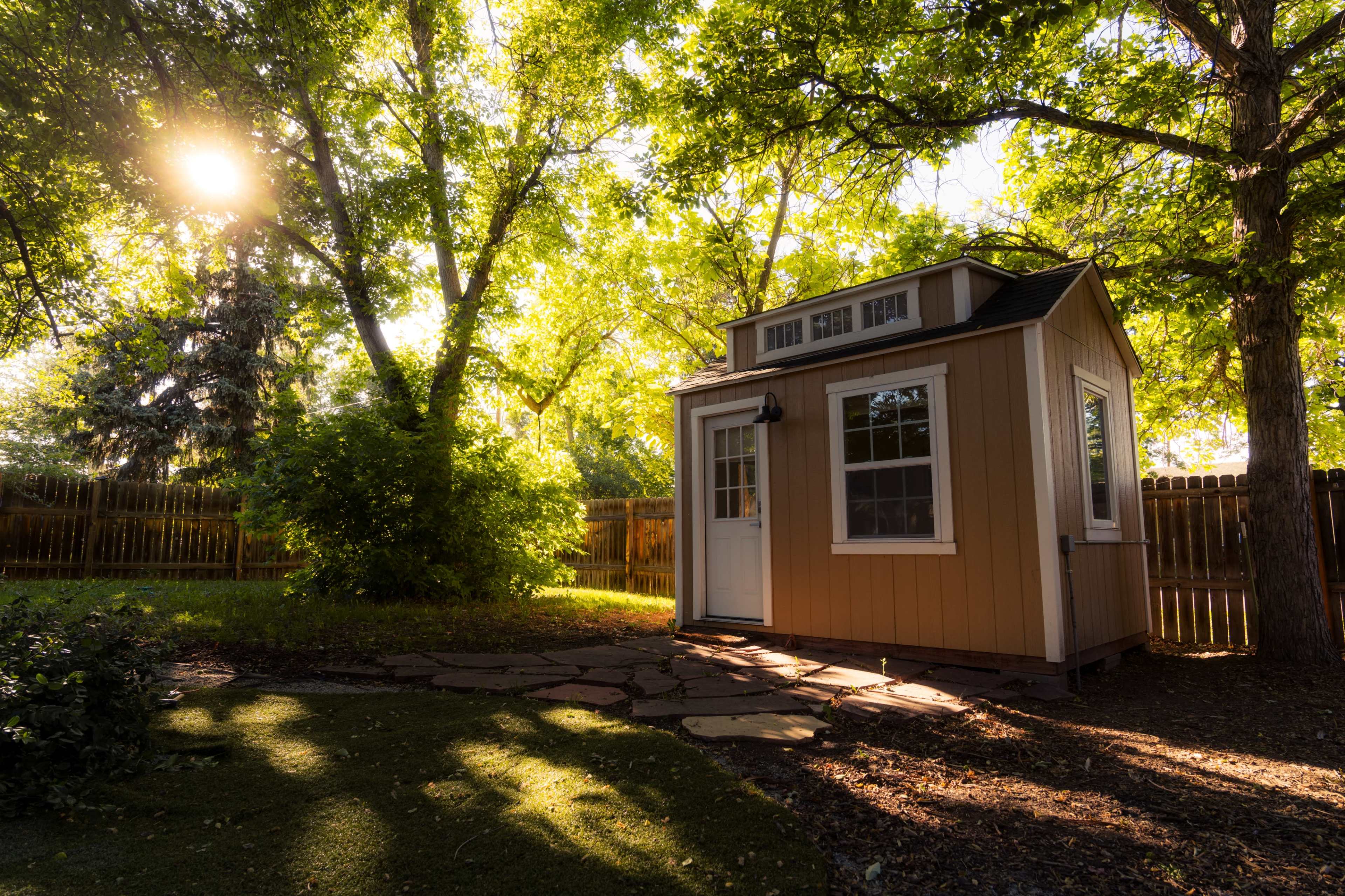 A small, wooden shed with white trim sits in a sunlit backyard surrounded by trees and a stone pathway.
