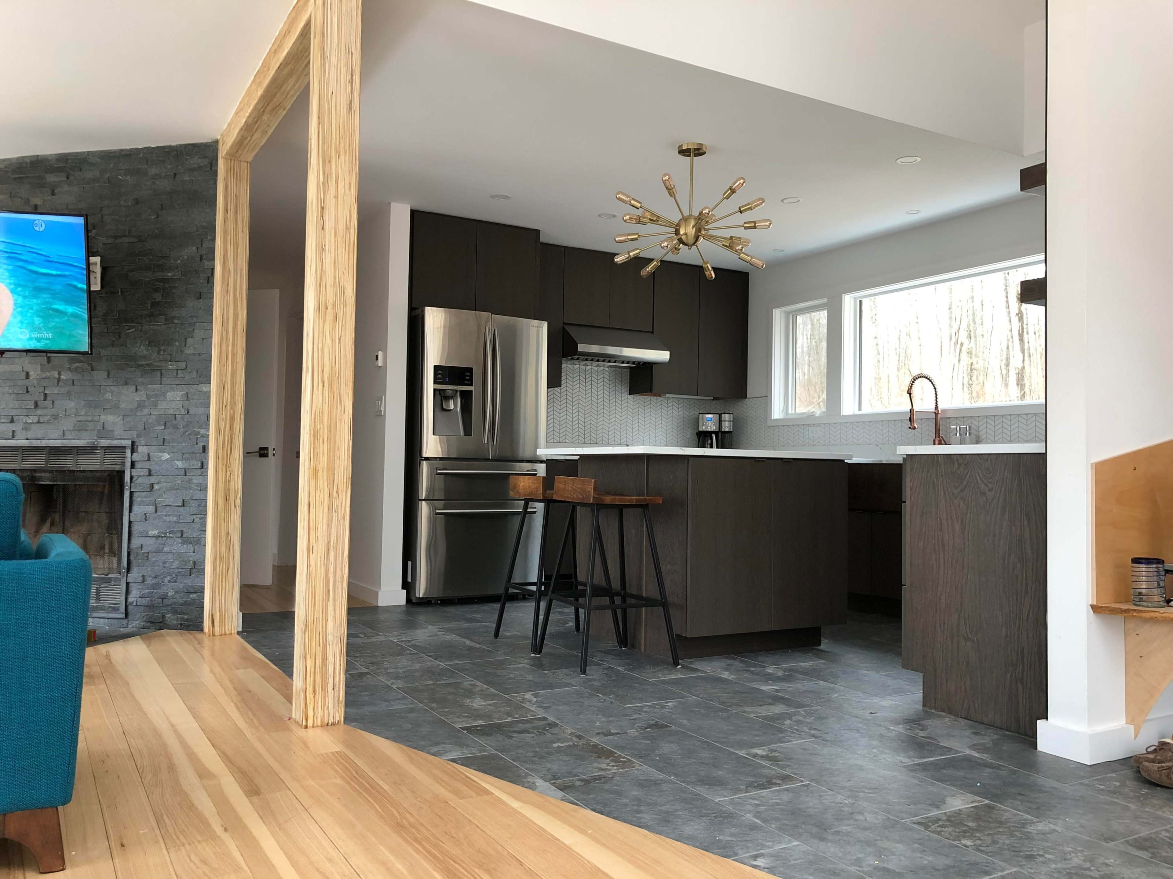 A modern kitchen features dark cabinetry, stainless steel appliances, and large windows, with an adjacent living area visible through a wooden frame.