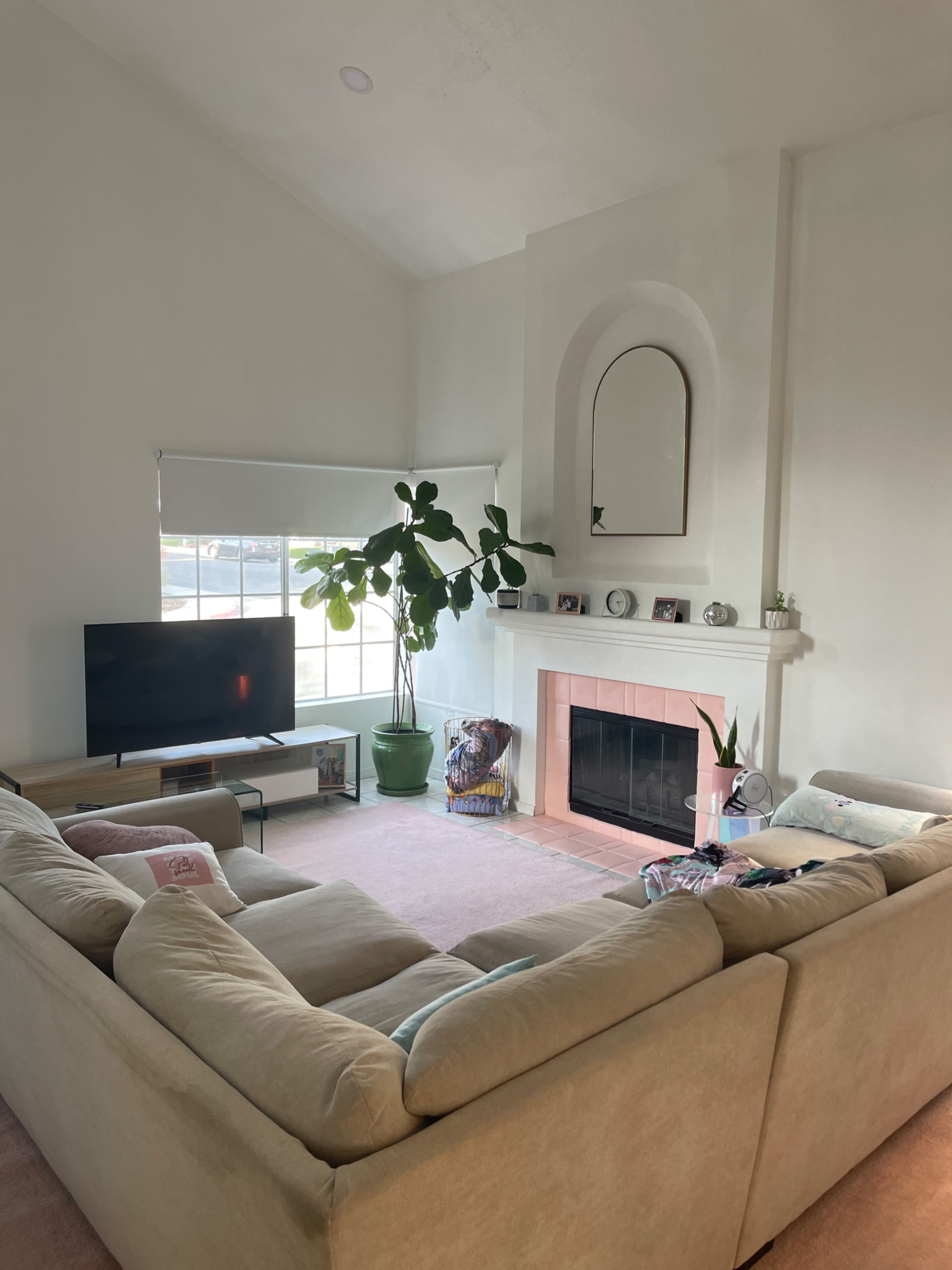 A spacious living room features a large sectional sofa, a television, a fireplace with a mirror above it, and a tall plant near a window.