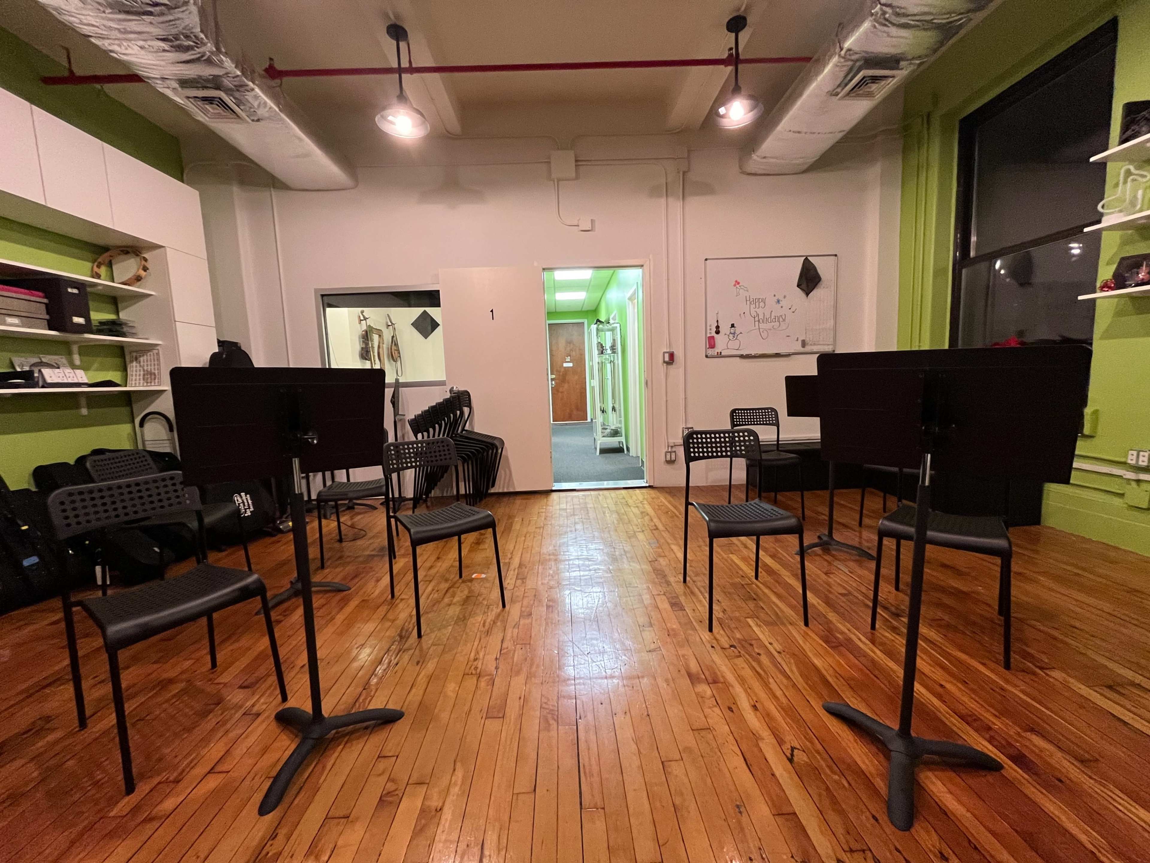 A music rehearsal room features several black music stands arranged in a circular formation on a wooden floor, with green walls and an open door leading to another area.