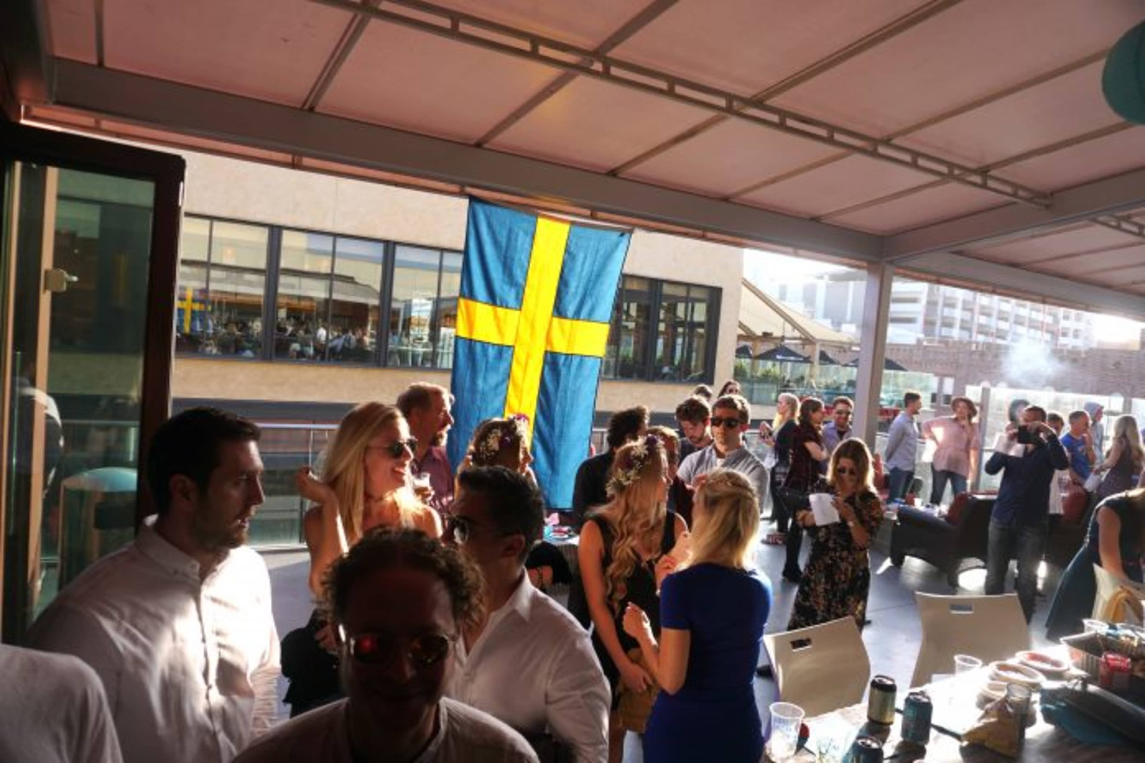 A crowd of people socializes under a Swedish flag in a lively outdoor setting.