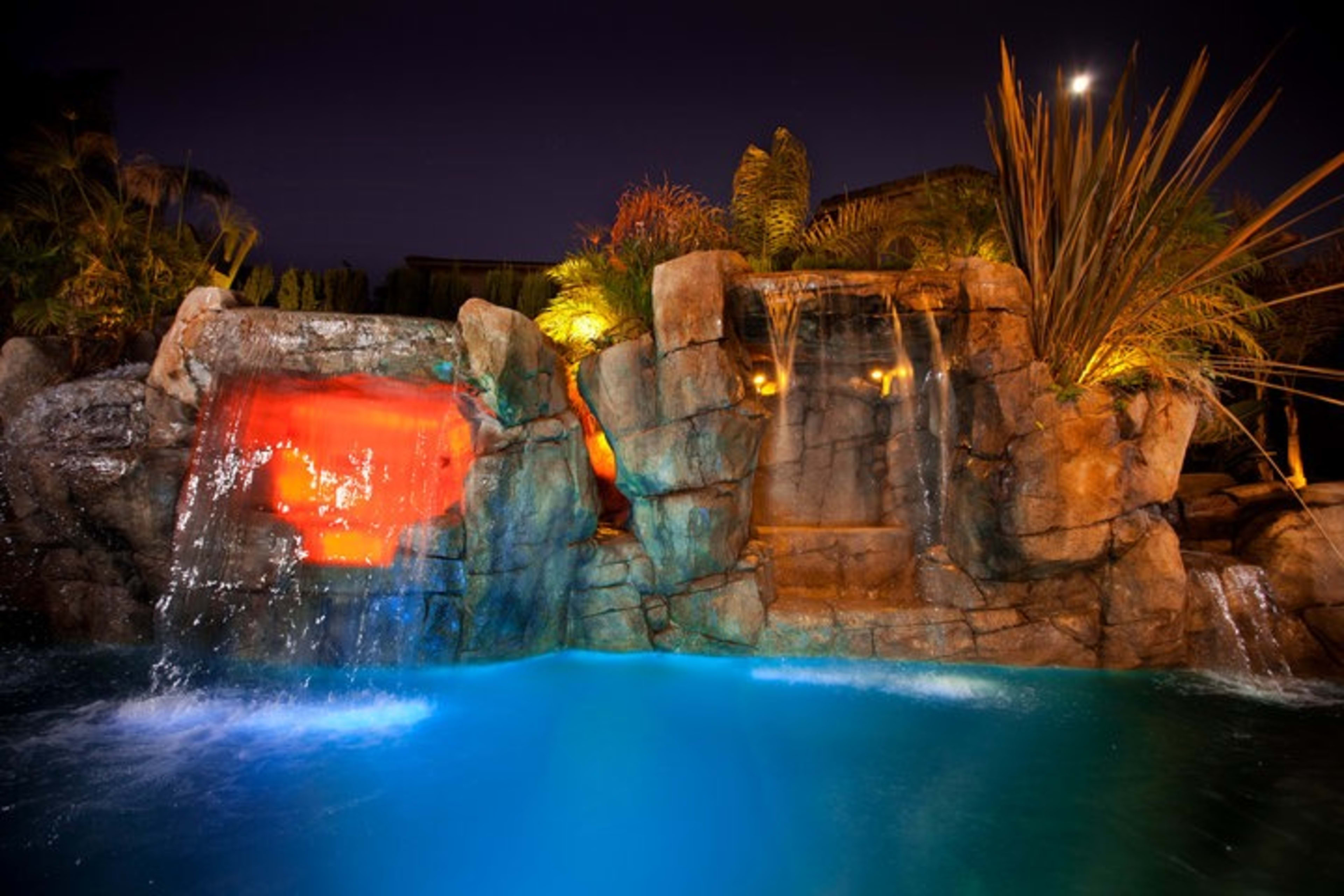 A brightly lit water feature cascades over rocky formations into a pool at night.