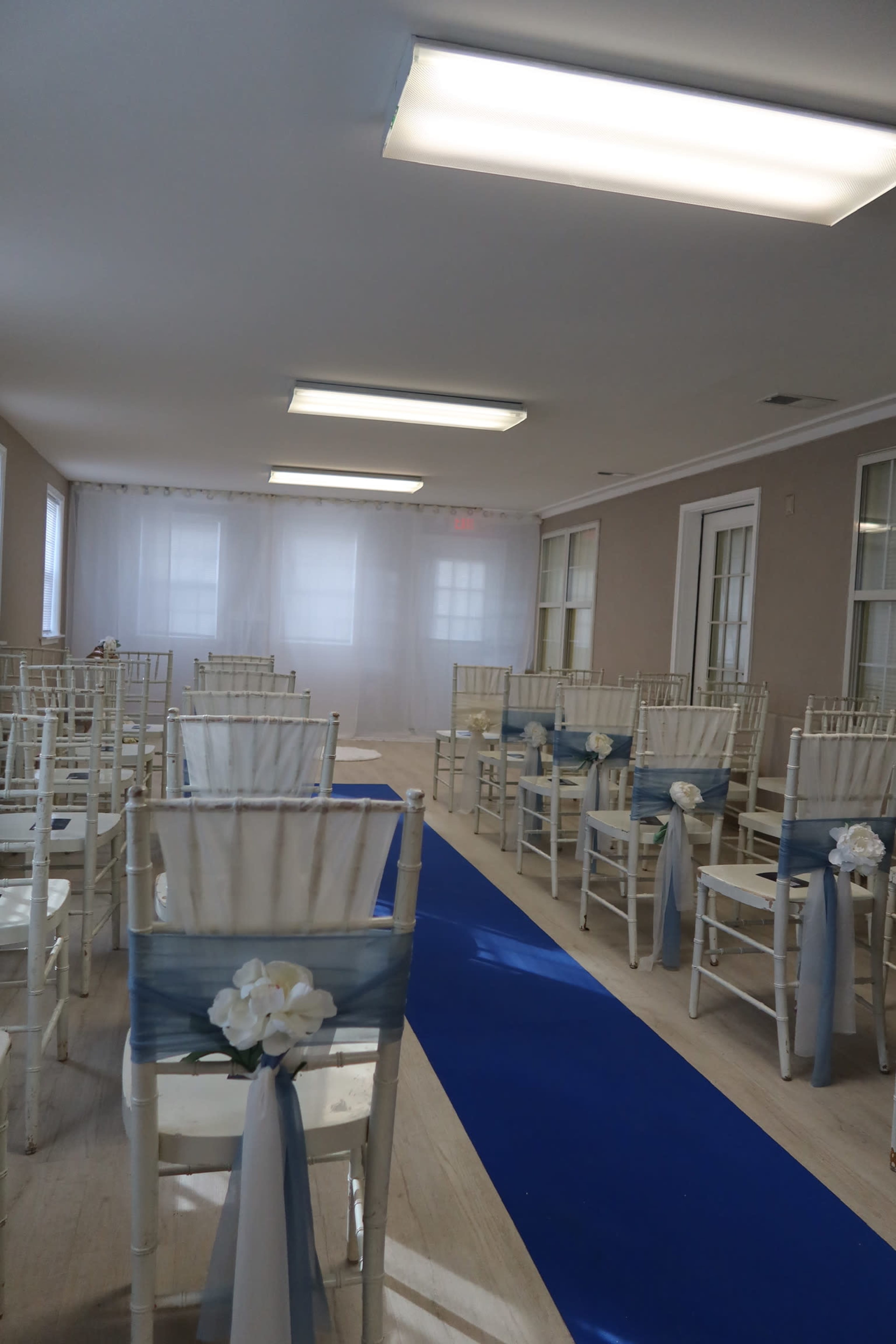 The image shows a wedding ceremony setup with white chairs adorned with blue sashes and flowers, arranged along a blue aisle runner.
