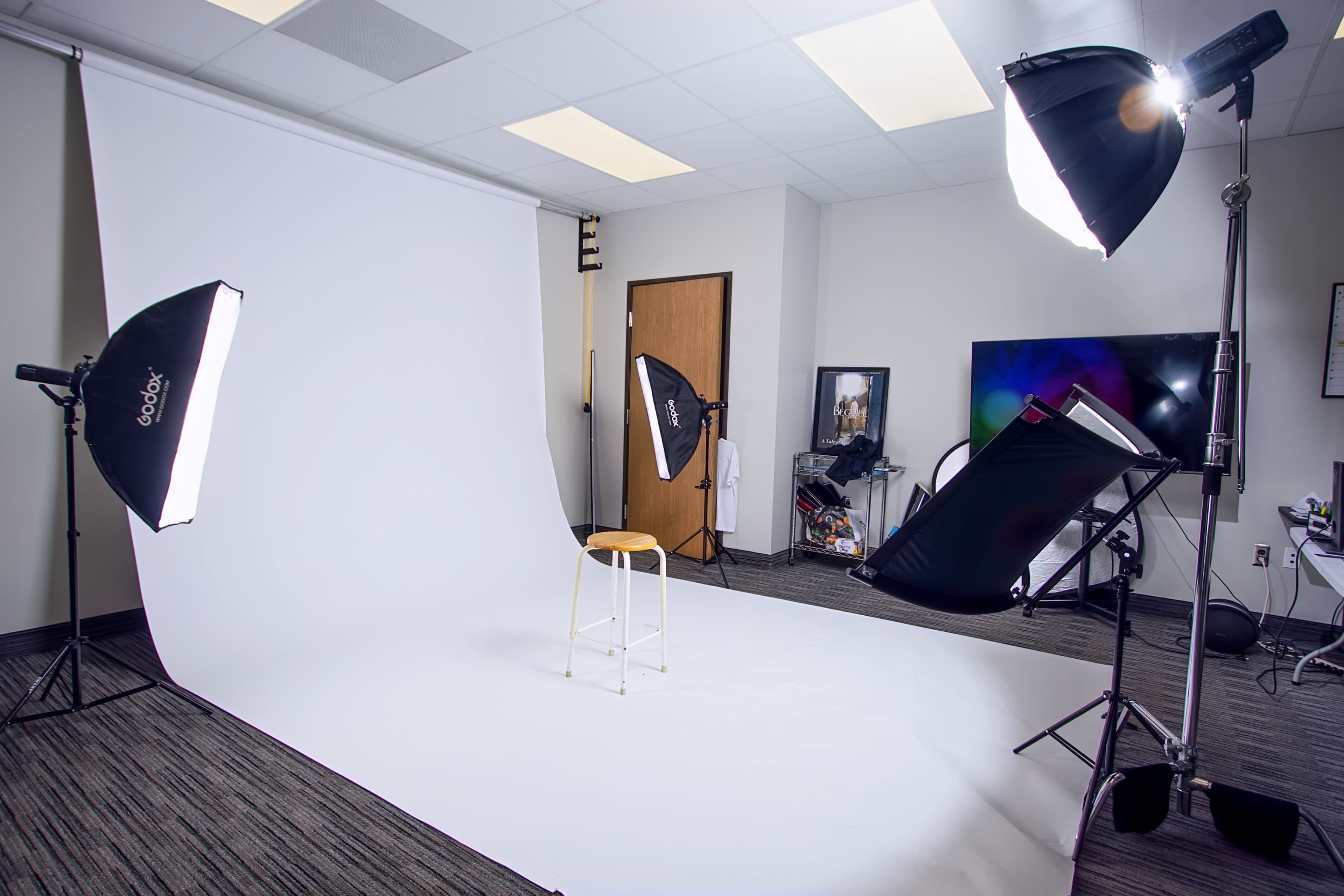The image shows a photography studio with a white backdrop, a wooden stool in the center, and multiple softbox lights positioned around the area.
