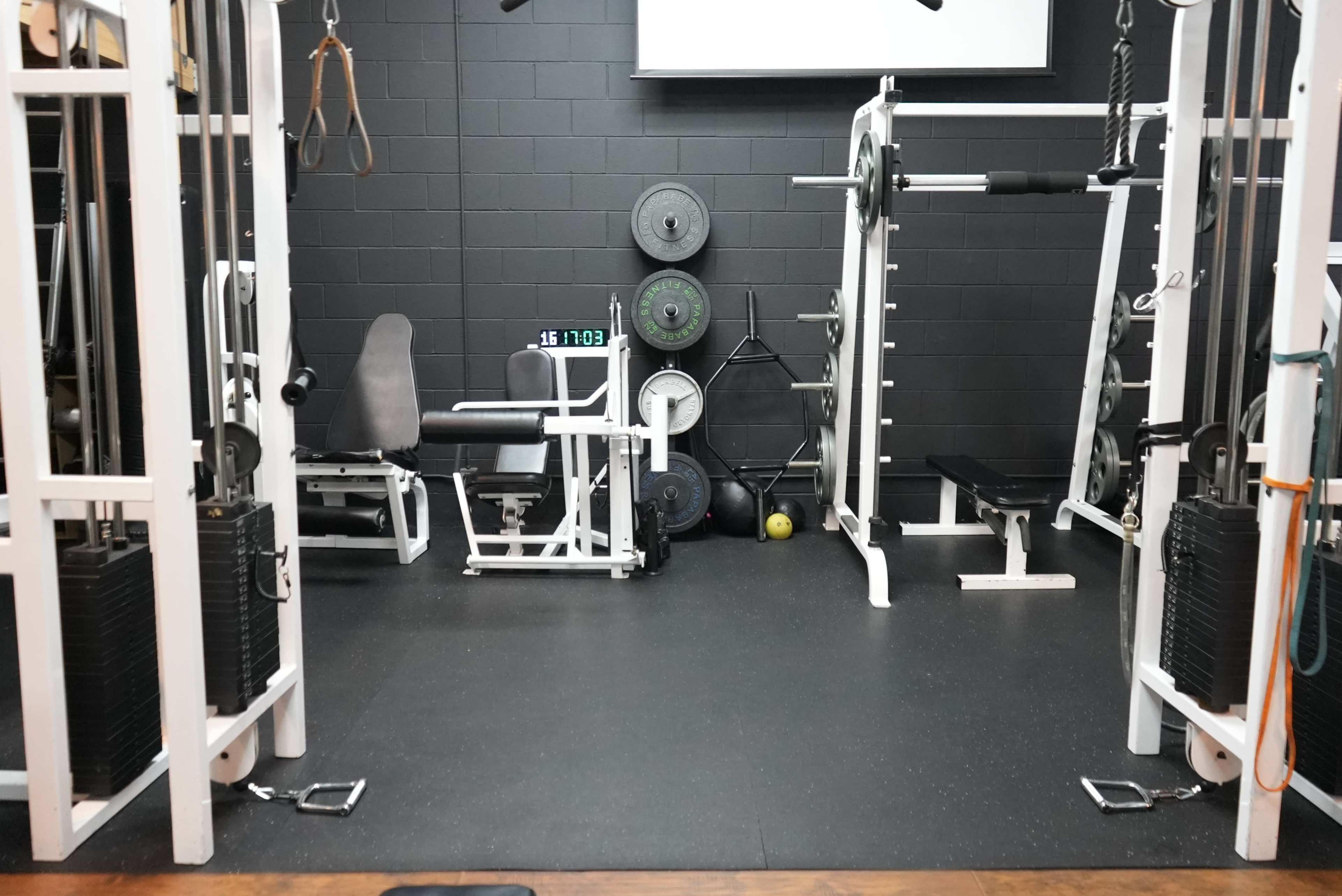 A gym interior with various weightlifting equipment, including a bench, barbell racks, and free weights against a black wall.