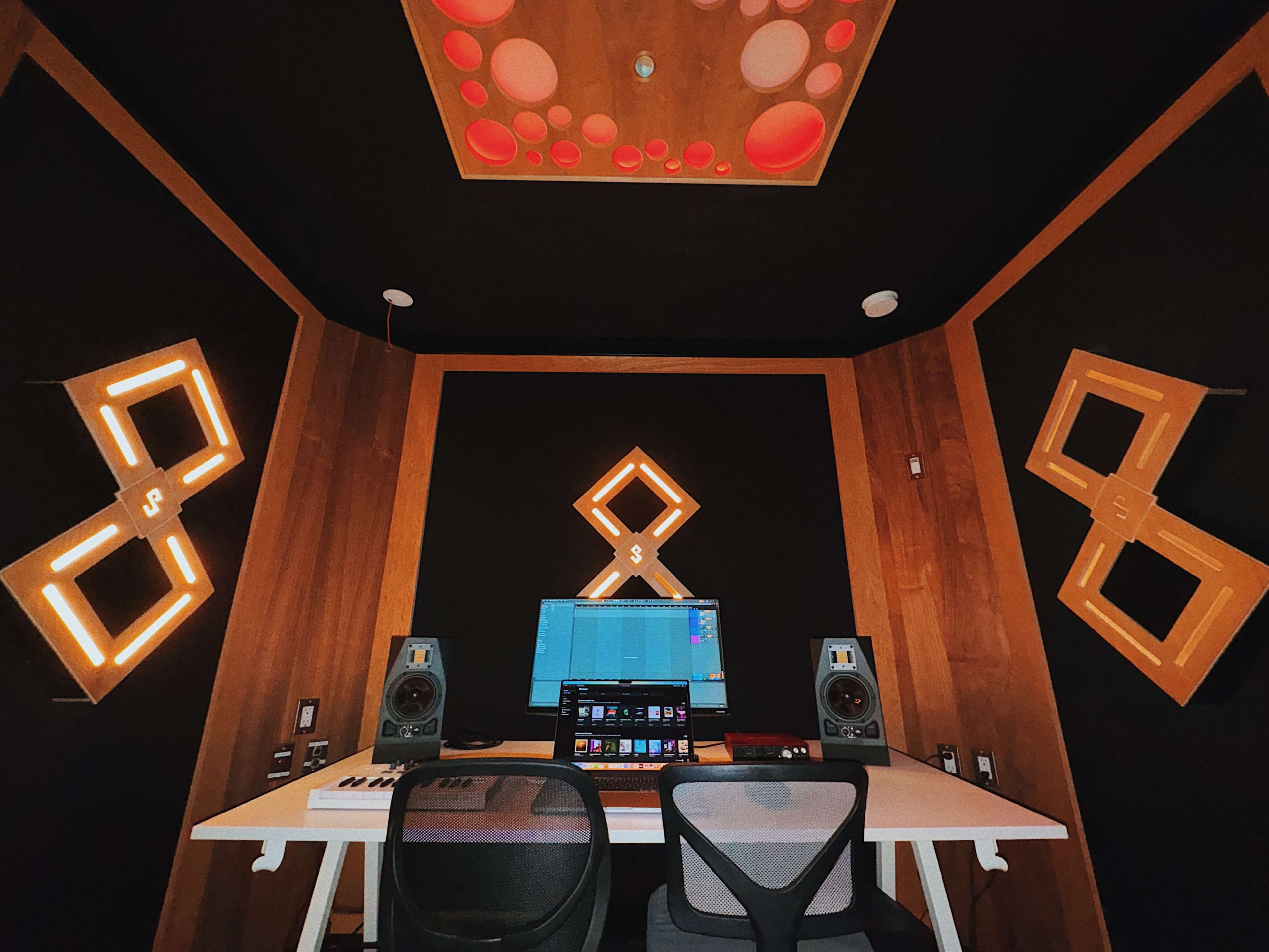 The image shows a modern music studio with a computer setup, two studio monitors, and illuminated wall designs on a black background.