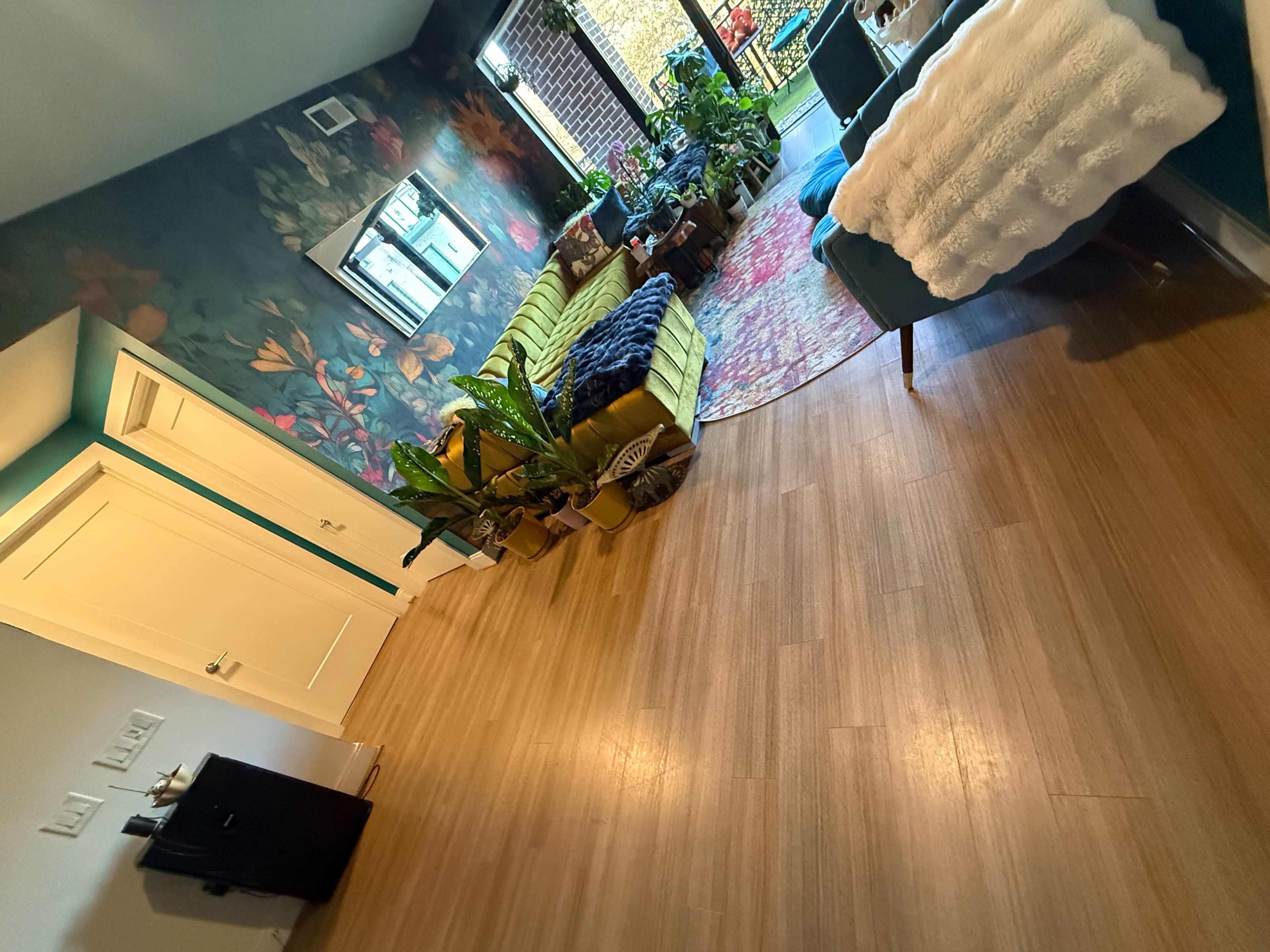 The image shows a stylish living room with a patterned wall, a green couch, various indoor plants, and a wooden floor.