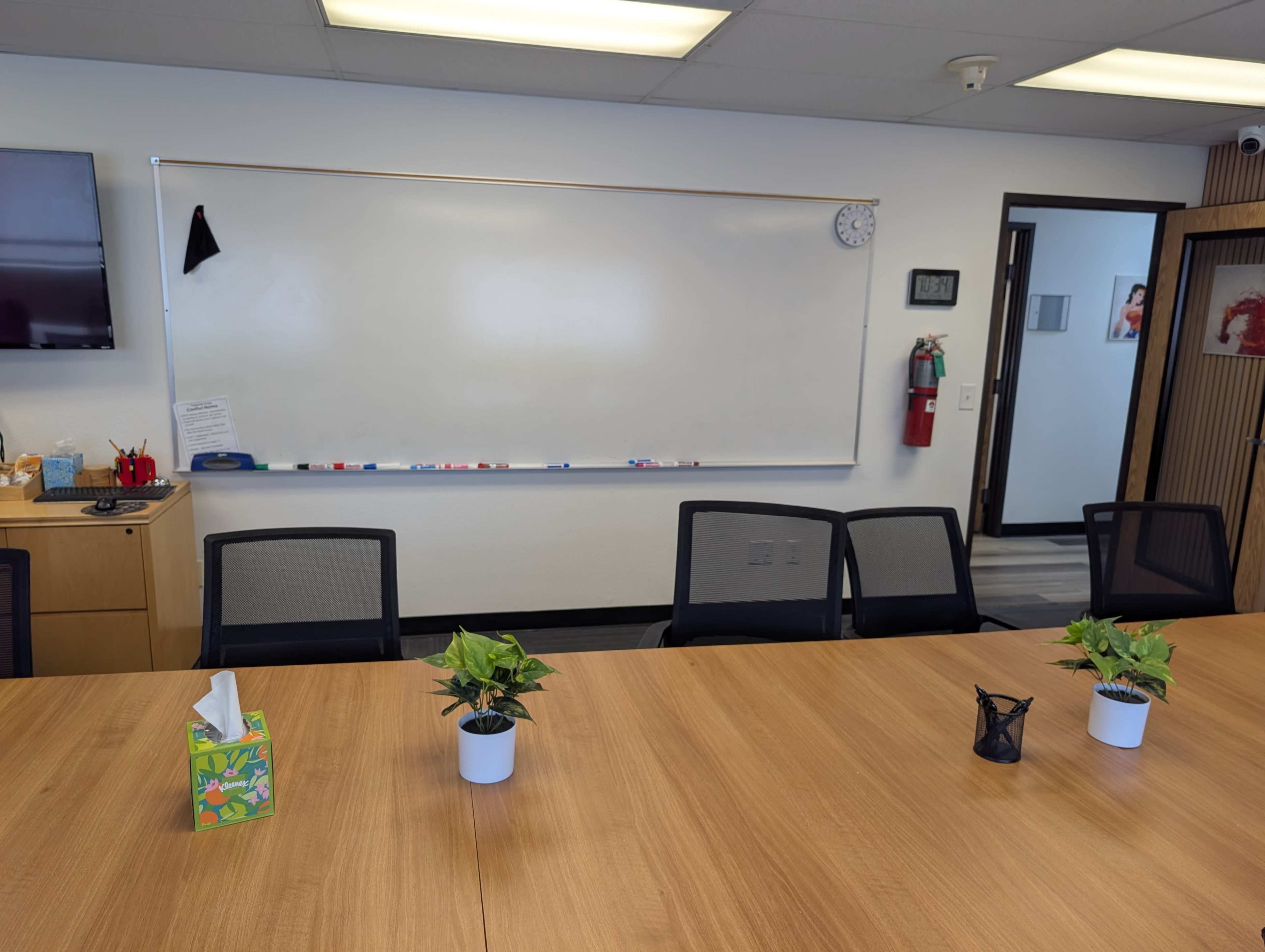 A conference room features a large wooden table surrounded by chairs, with small potted plants on the table and a whiteboard on the wall.