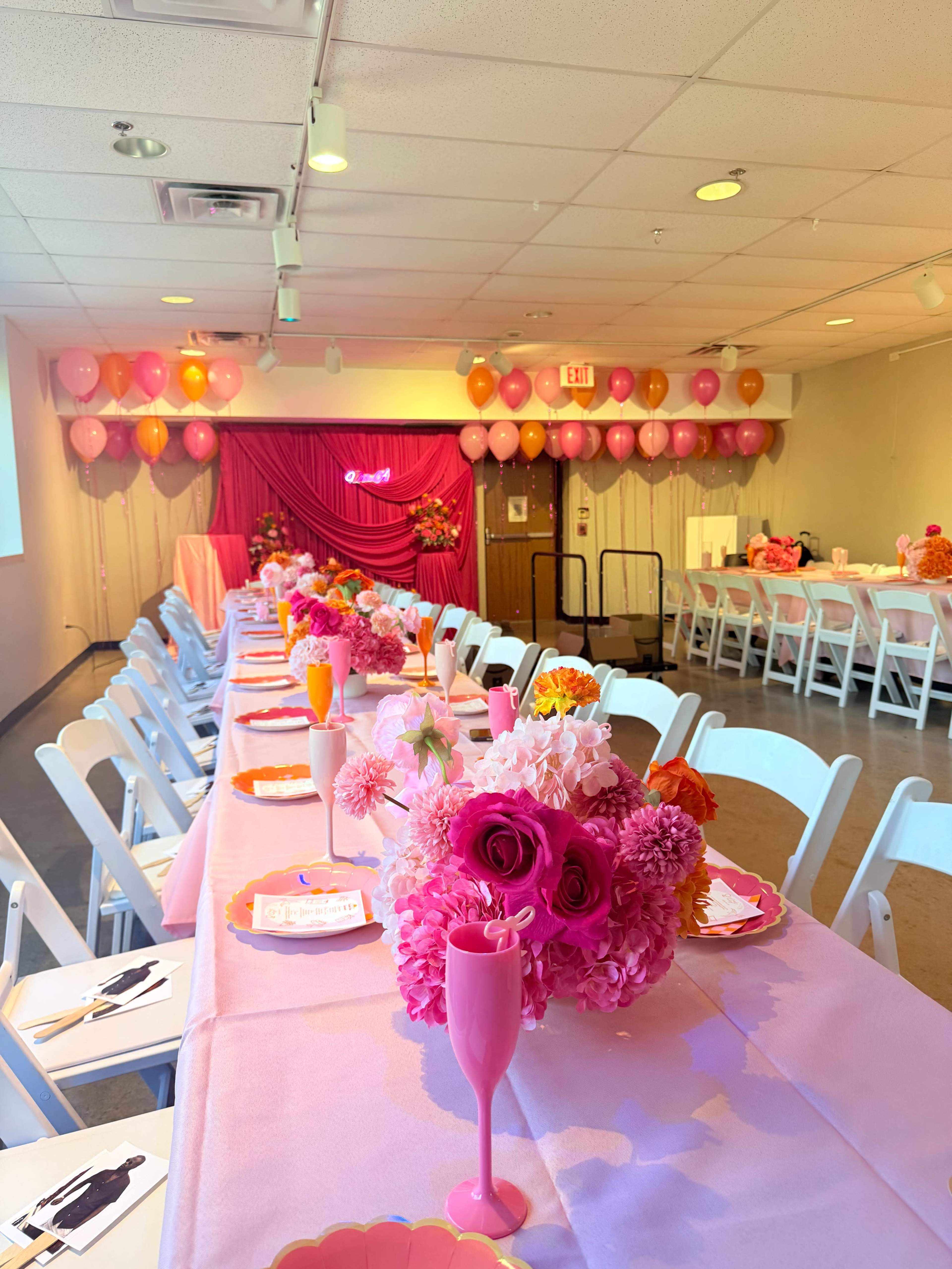 The image shows a decorated event space with long tables covered in pink tablecloths, adorned with floral centerpieces, pink balloons, and a backdrop for a celebration.
