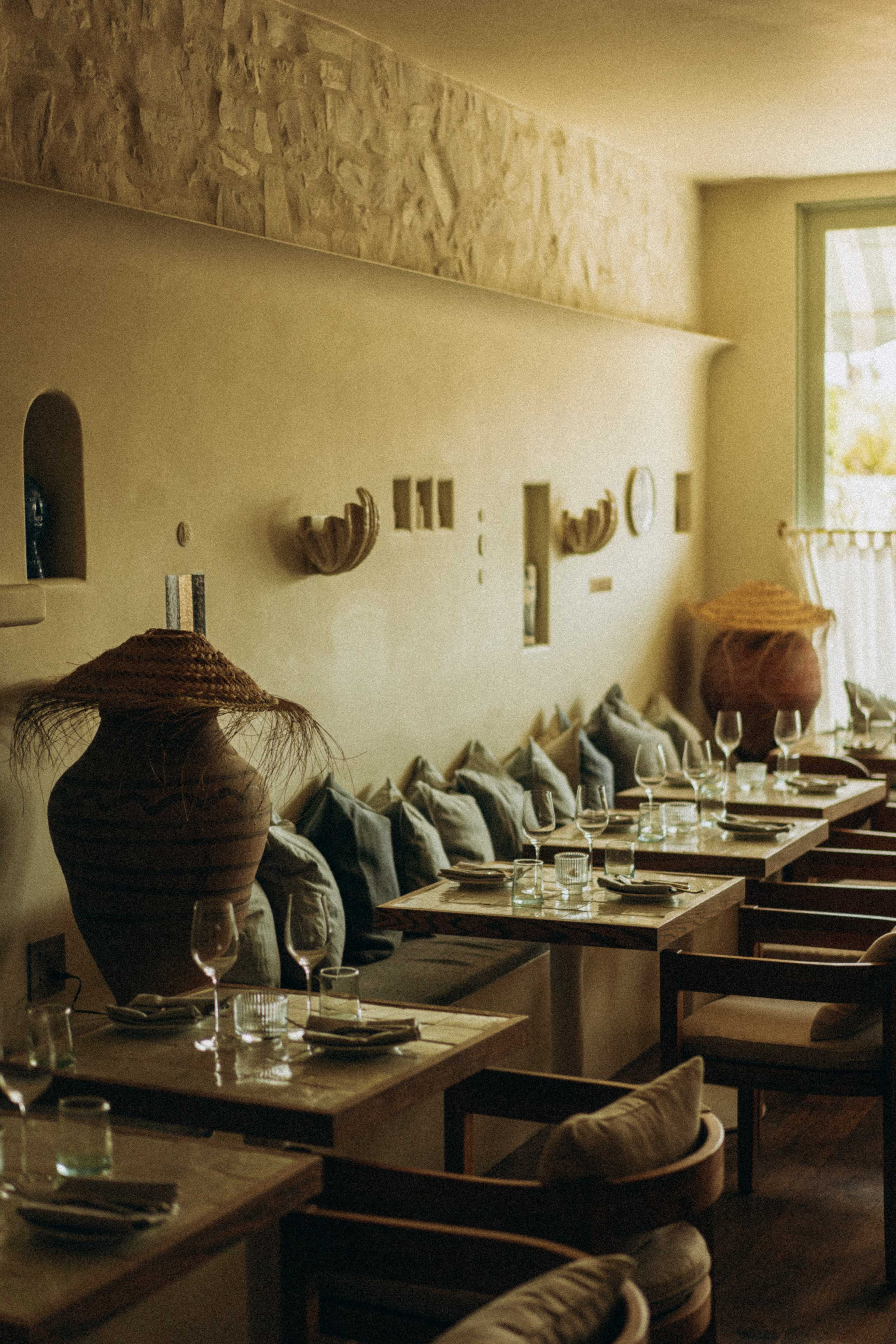 The image shows a cozy restaurant interior with wooden tables set with glassware and neatly arranged cushions along the walls.
