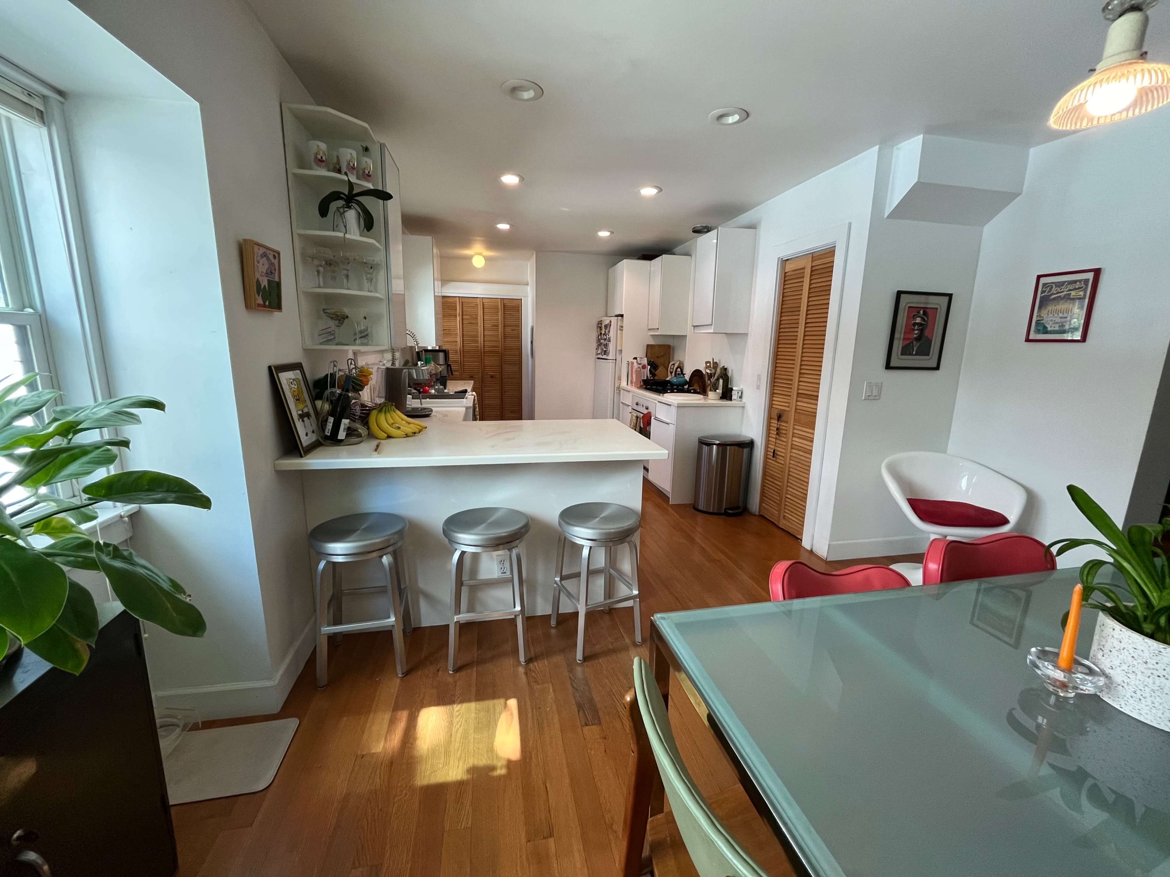 A modern kitchen features a white bar counter with metal stools, surrounded by cabinets and appliances, alongside a dining area with a glass table and chairs.