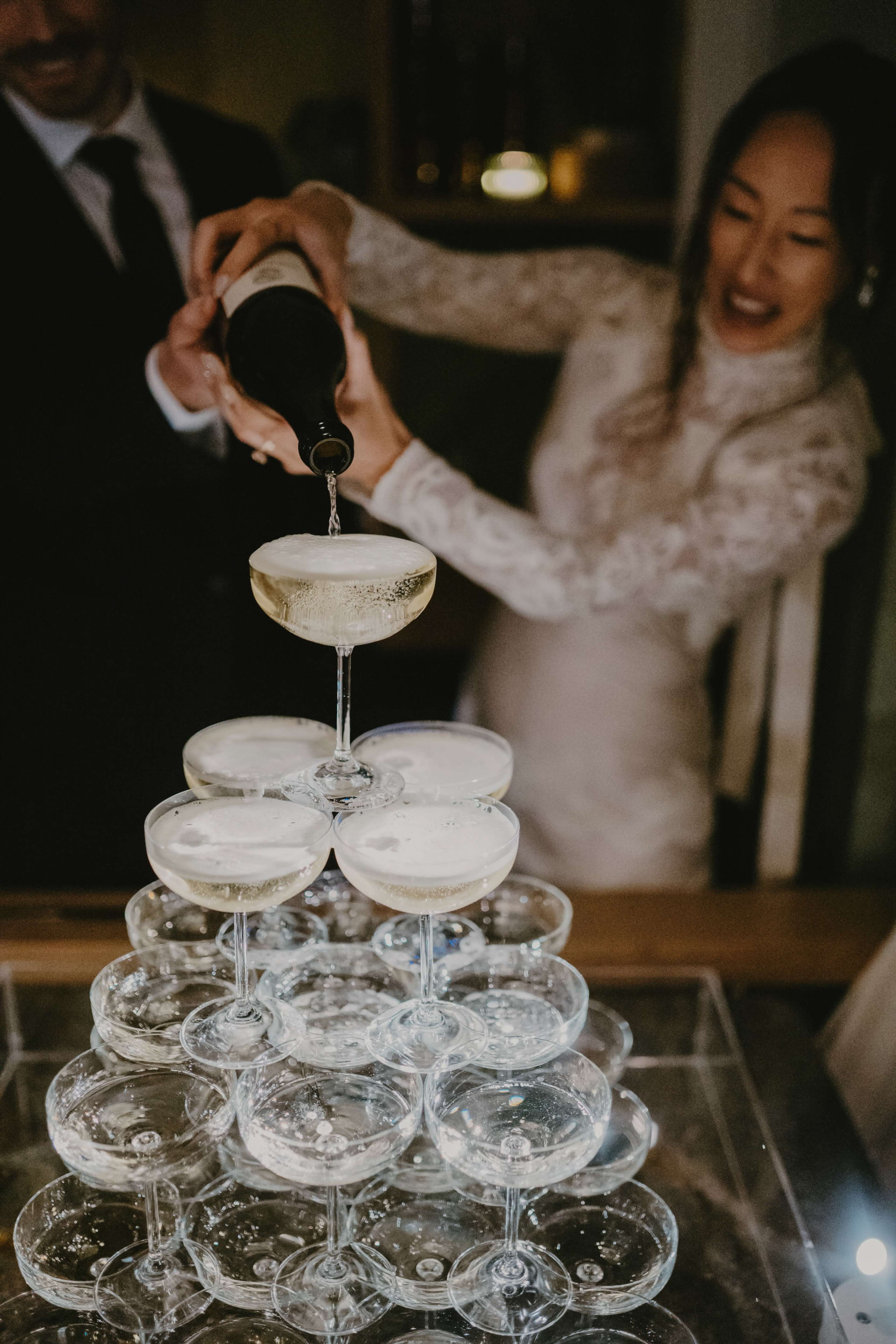 A person pours champagne into a pyramid of elegantly stacked glass coupe glasses at a celebration.