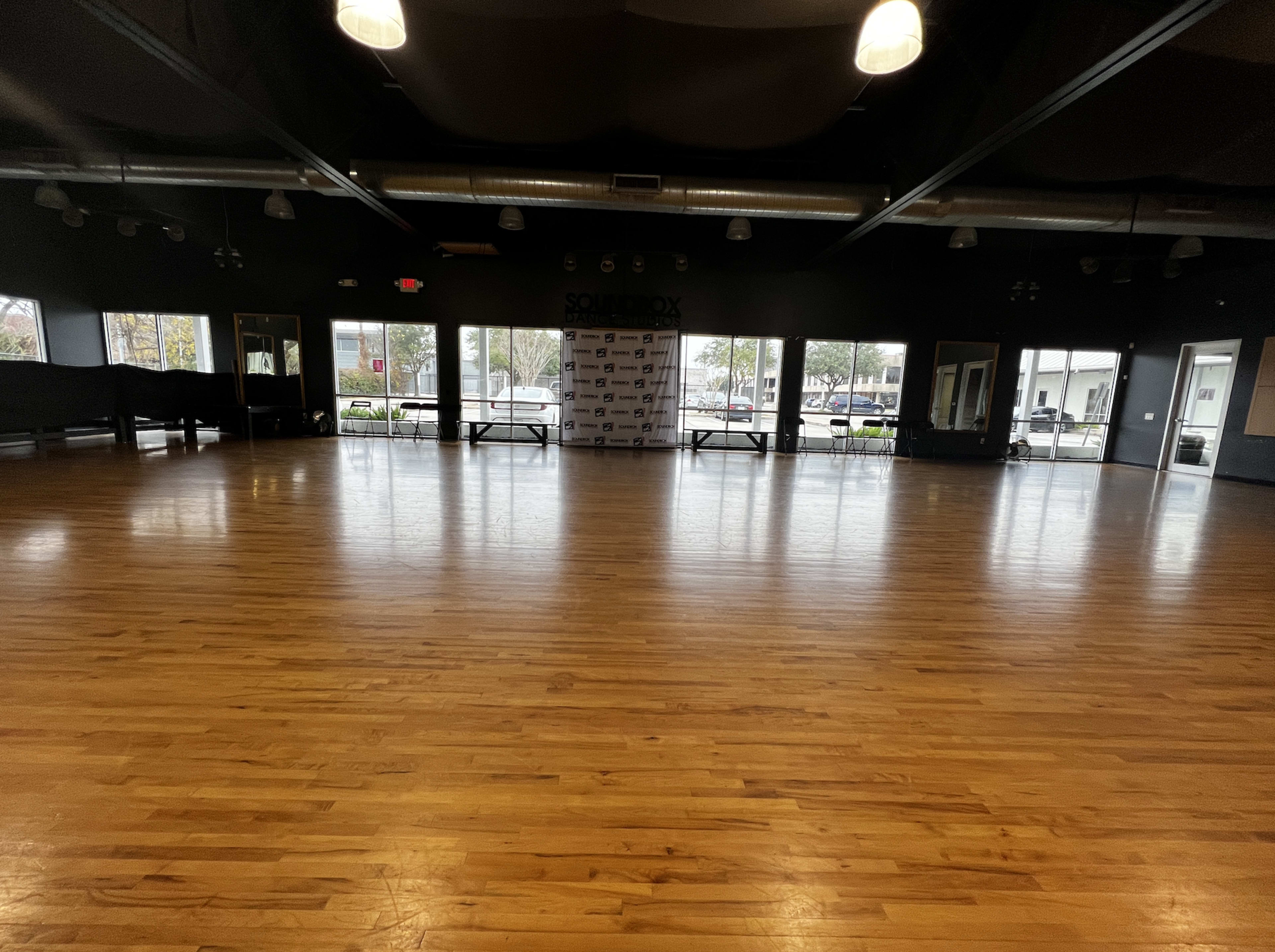 A spacious wooden dance studio with large windows, a branded backdrop at the front, and empty floor space.