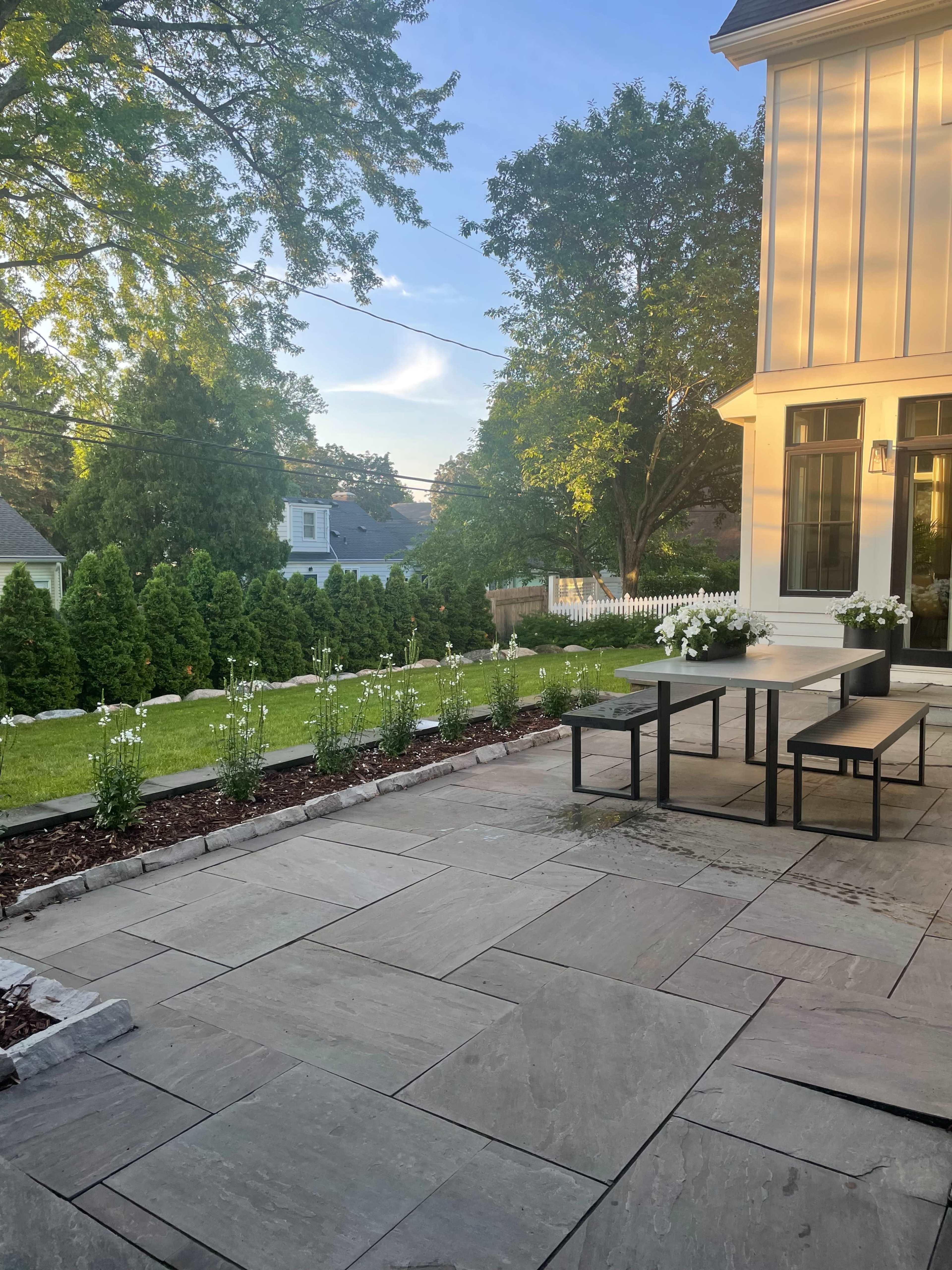 A patio area with a stone surface, a table and bench set, and neatly arranged flower beds and shrubs in the background.
