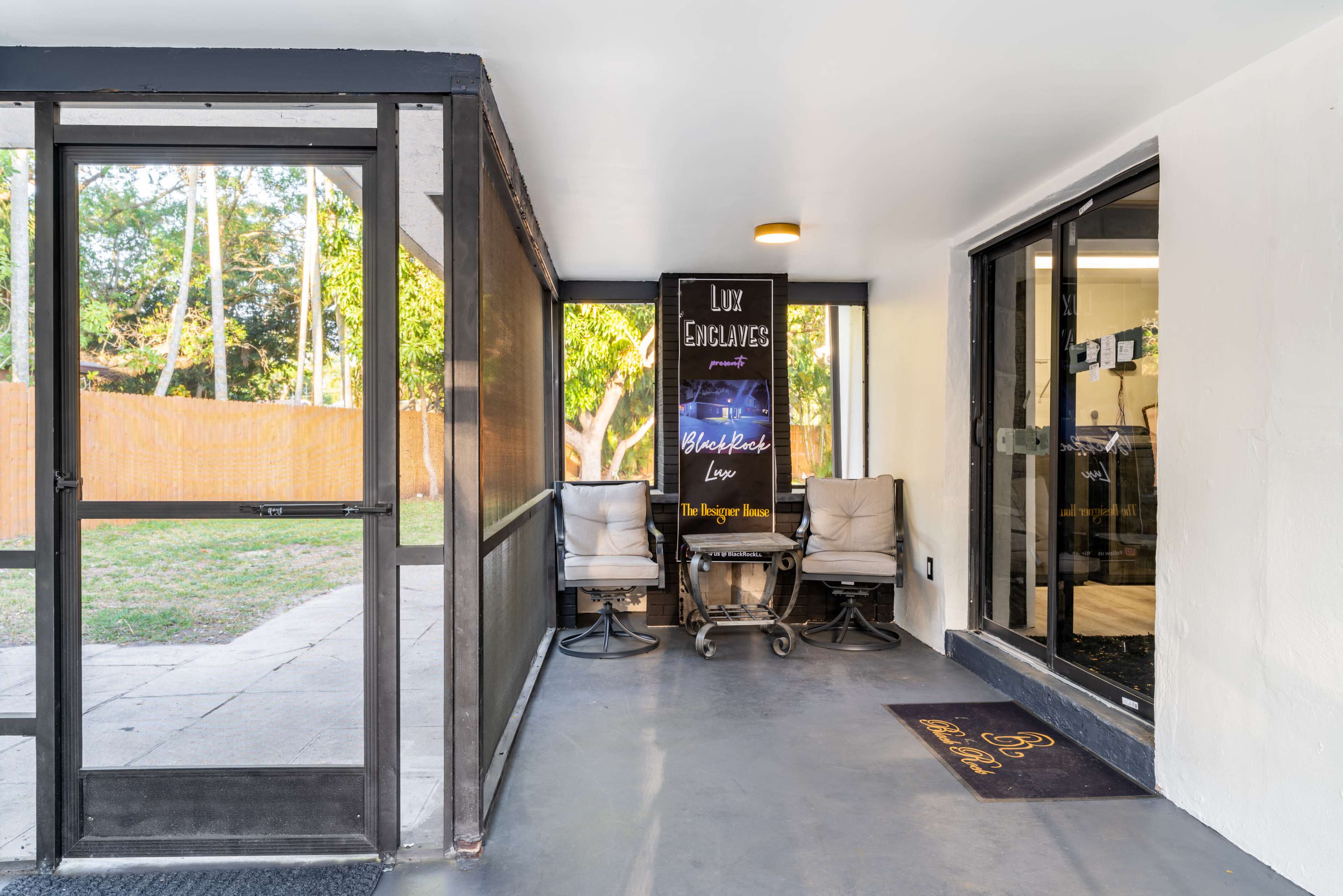 The image shows a screened porch area with two chairs and a sign displaying "Lux Embroideries" along with an entrance to an indoor space.