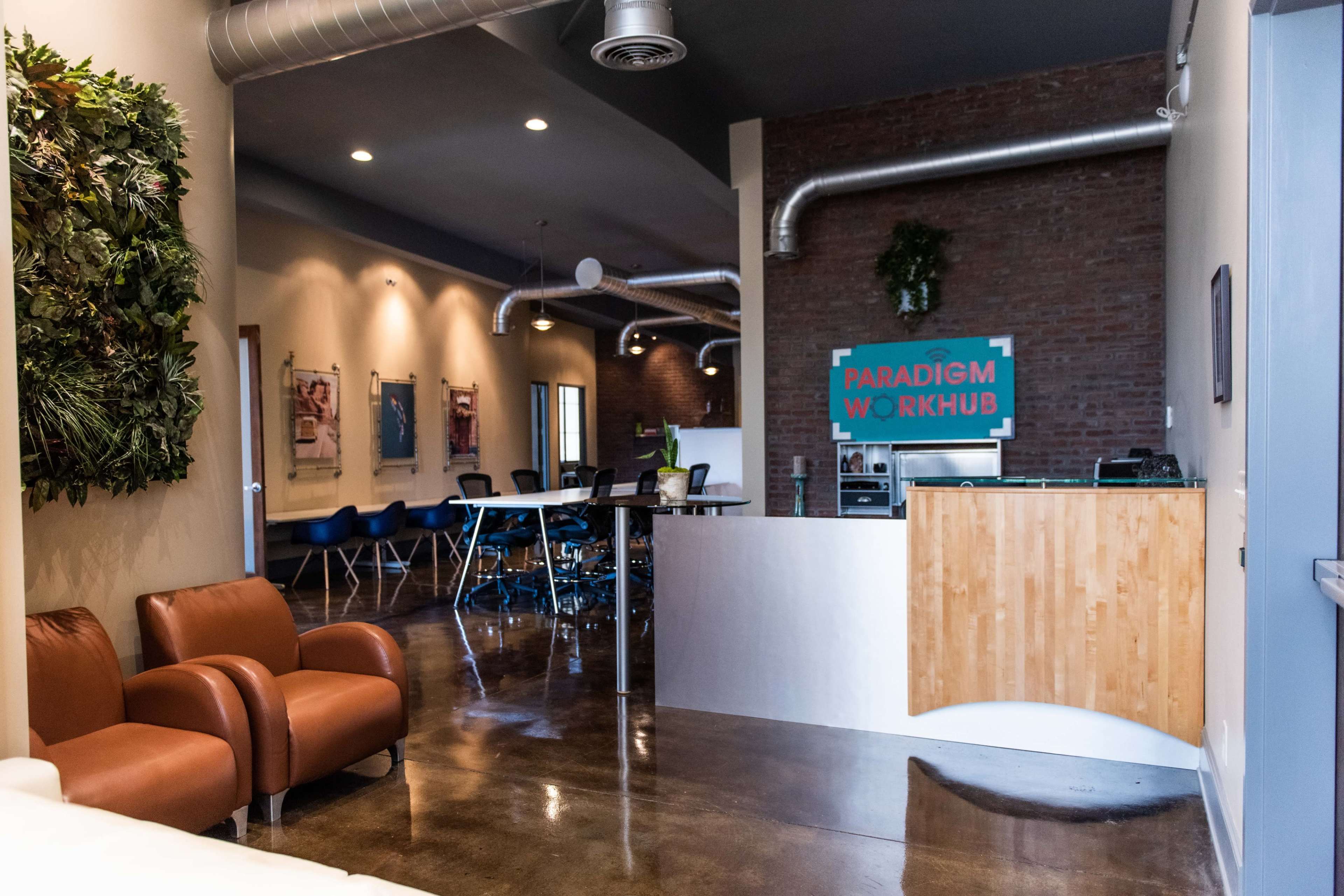 A modern office space featuring a reception area with a wooden desk, brown chairs, and a meeting area with tables and chairs, set against a backdrop of exposed brick walls.