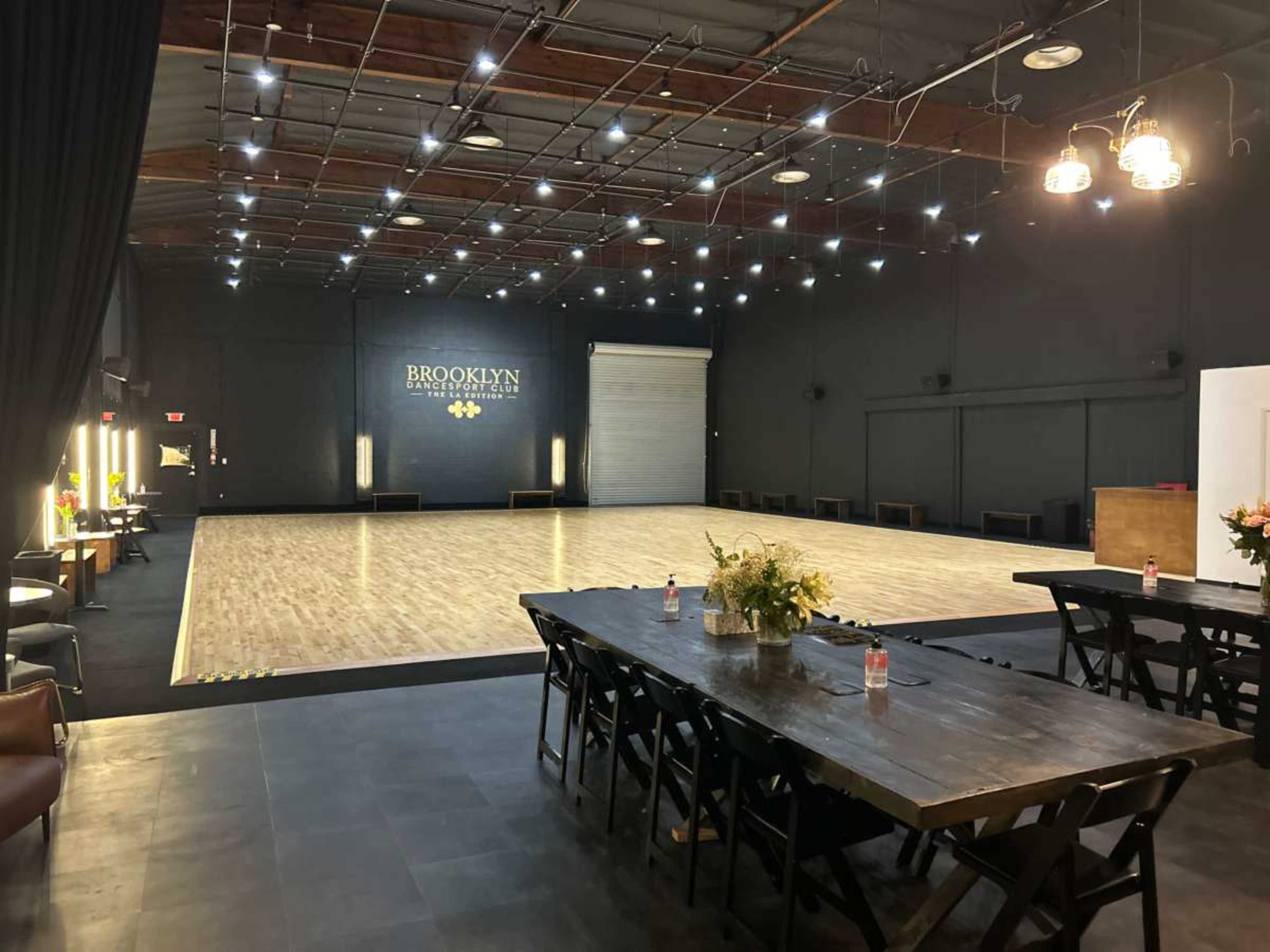 The image shows a spacious dance studio with a polished wooden floor, surrounded by black walls and a long table set for an event.