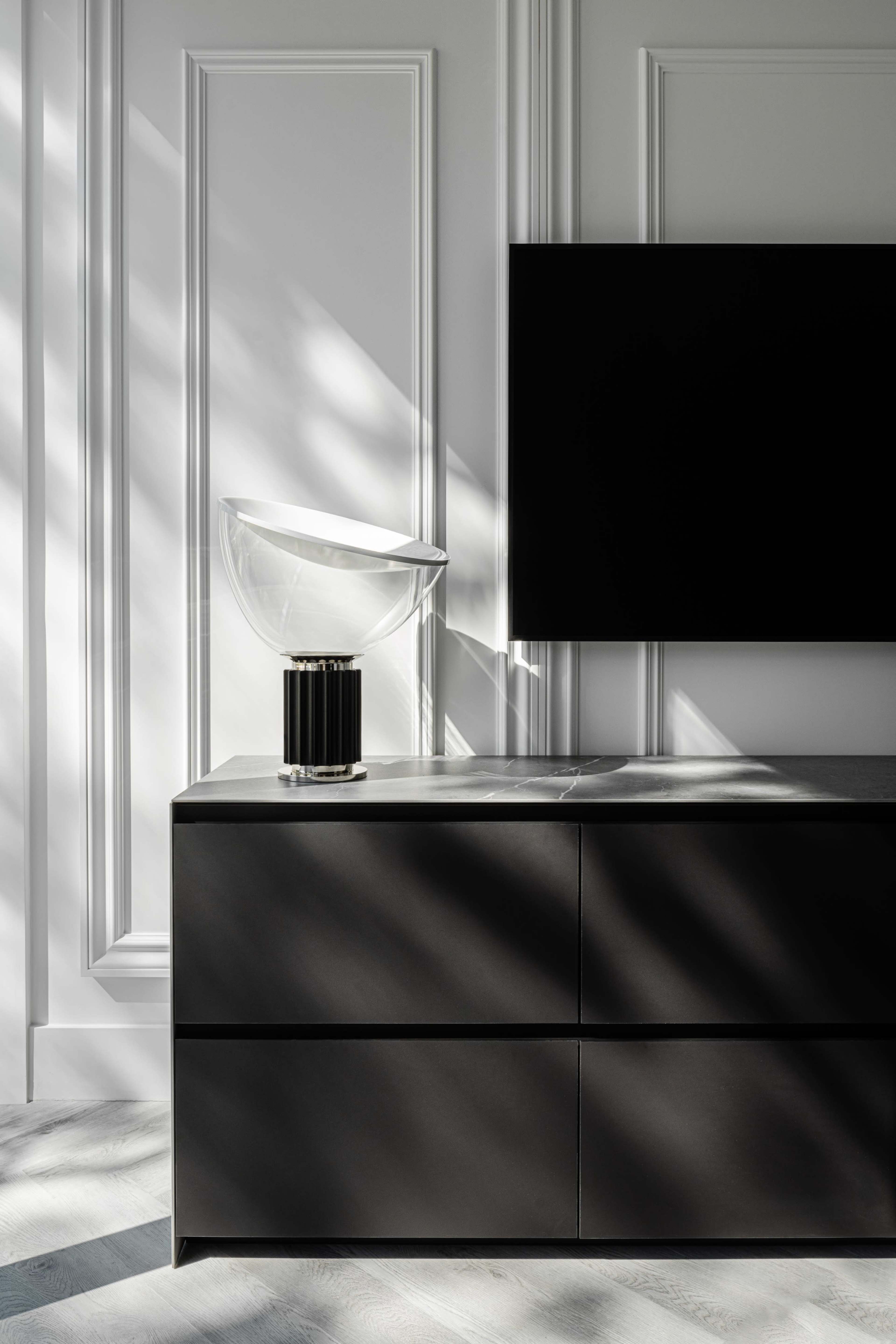 A sleek black cabinet with four drawers and a clear decorative lamp sits beneath a flat-screen TV against a wall featuring paneled molding and dramatic shadow play.