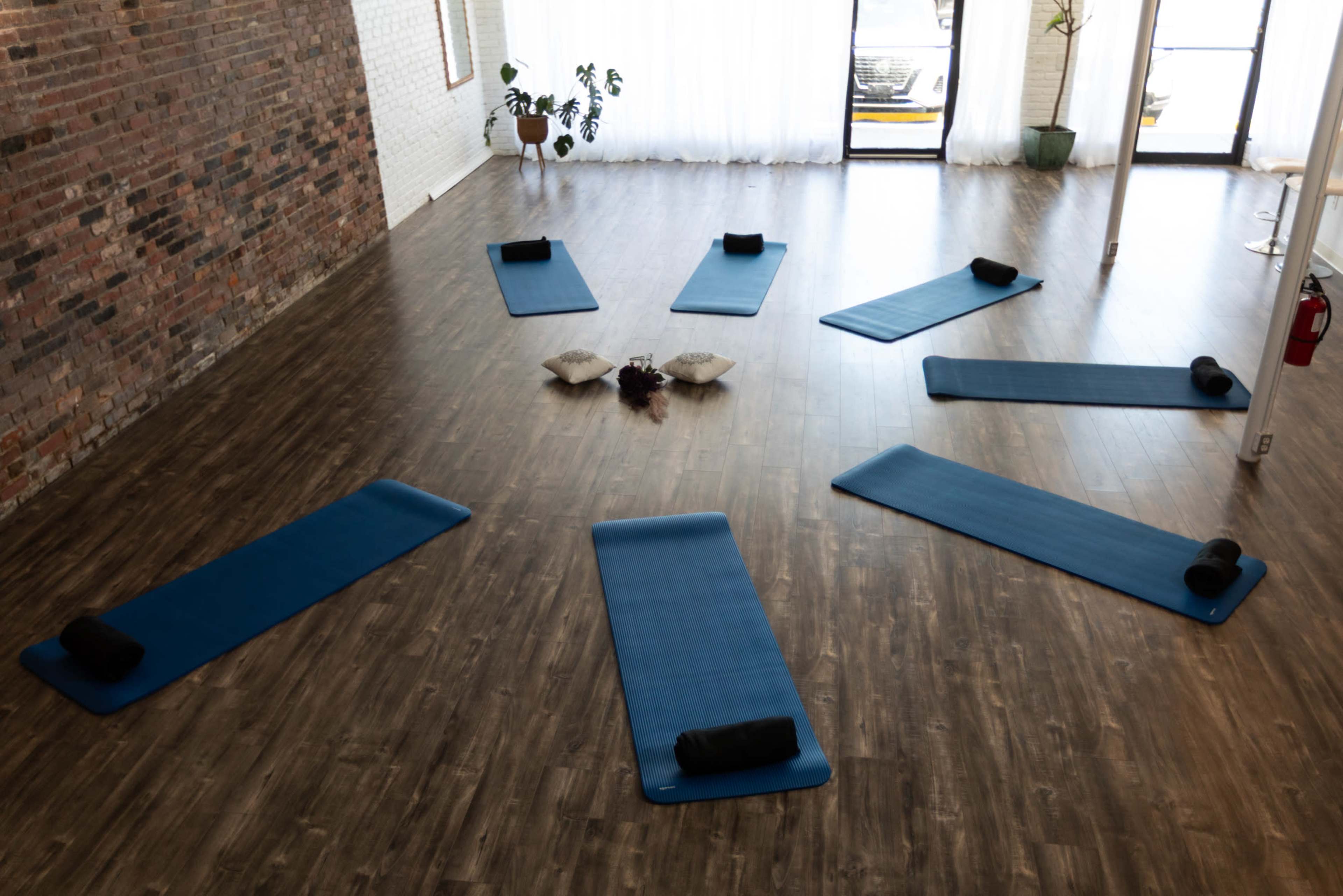 The image shows an airy yoga studio with several blue mats arranged in a circular pattern on a wooden floor, surrounded by plants and decorative cushions.