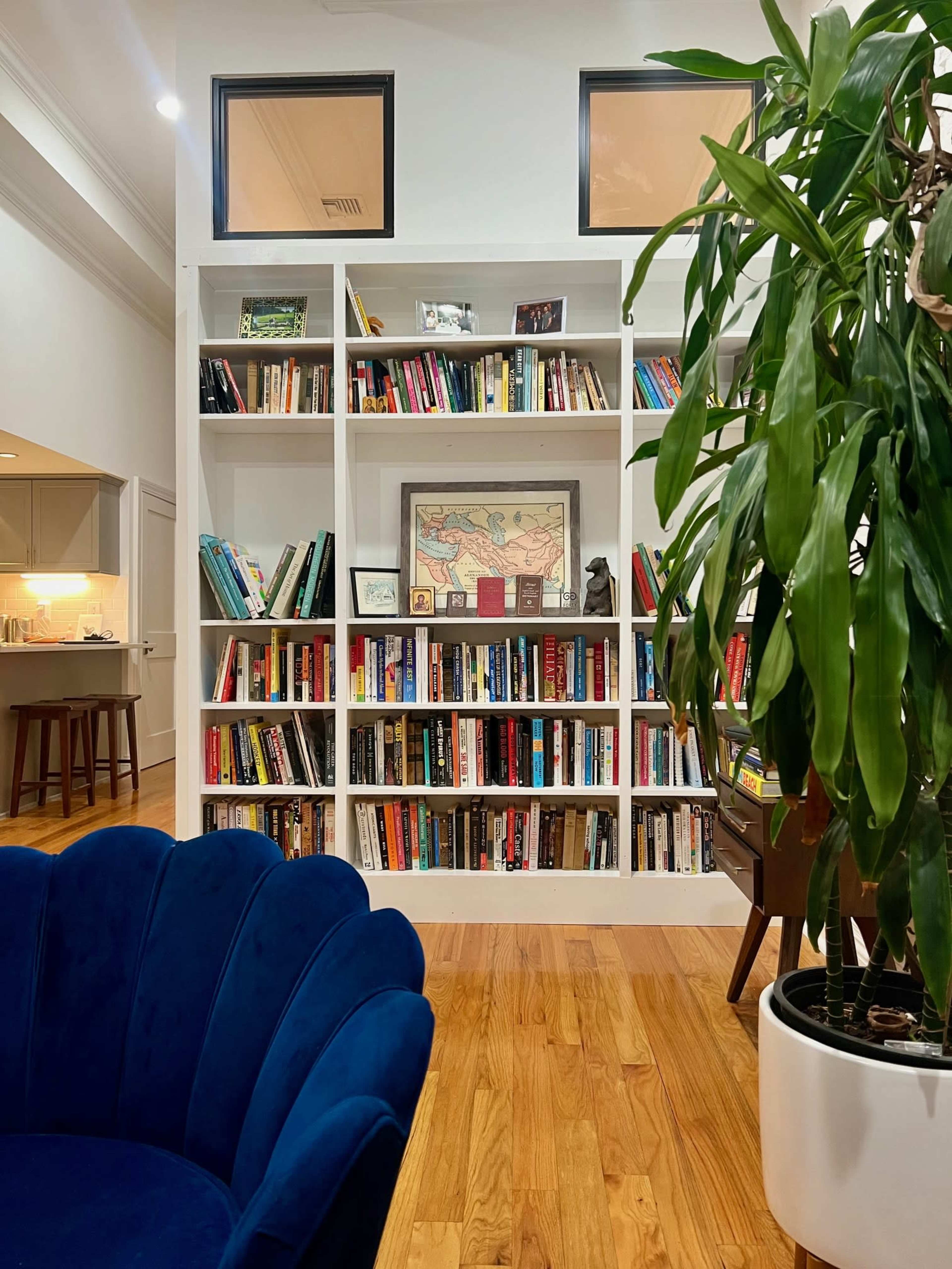 A bookshelf filled with various books is positioned next to a potted plant and a blue upholstered chair within a room that features large windows above the shelves.