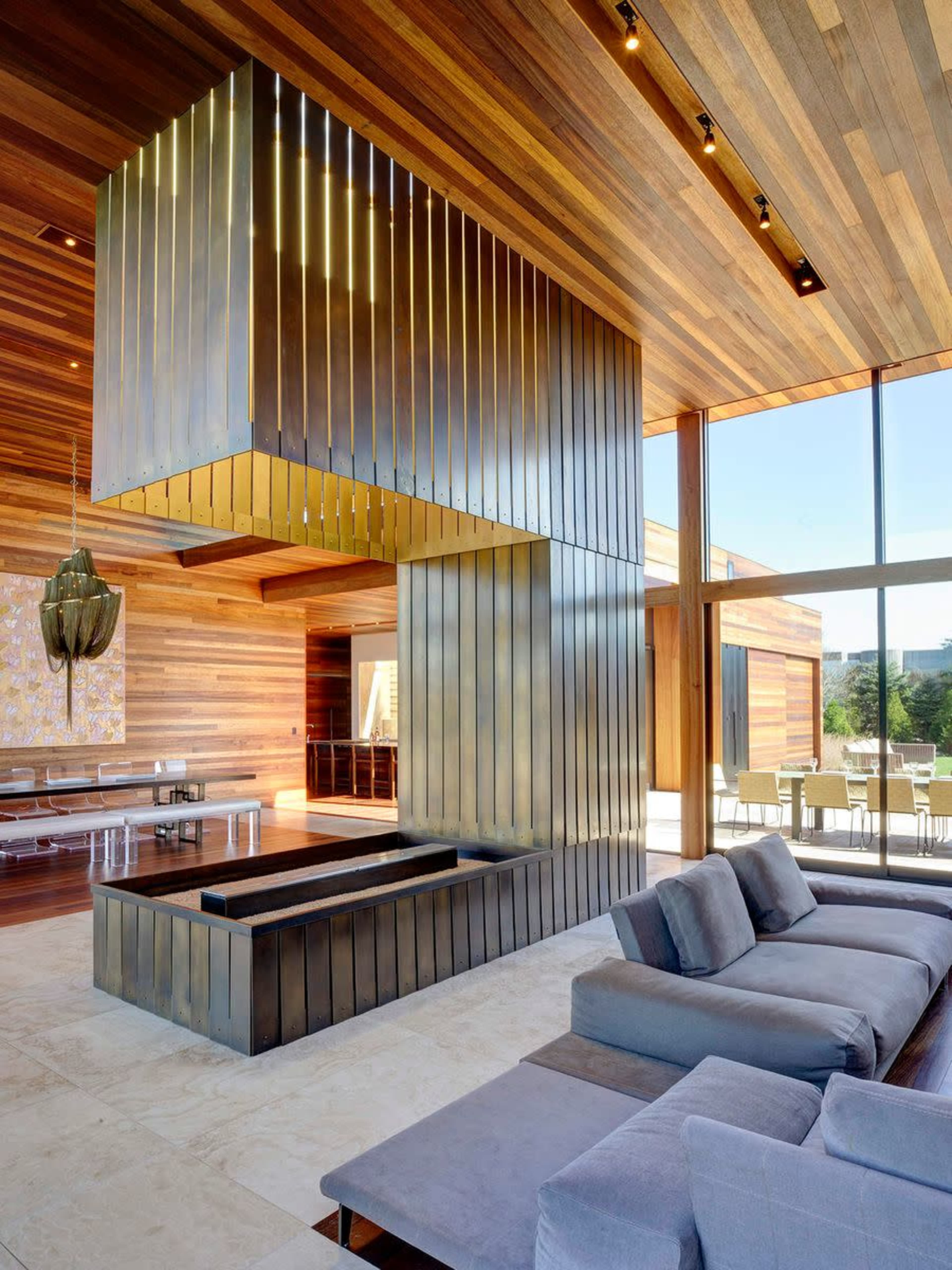 A modern living room featuring a metallic fireplace in the center, surrounded by wooden walls and large windows that allow natural light to enter.
