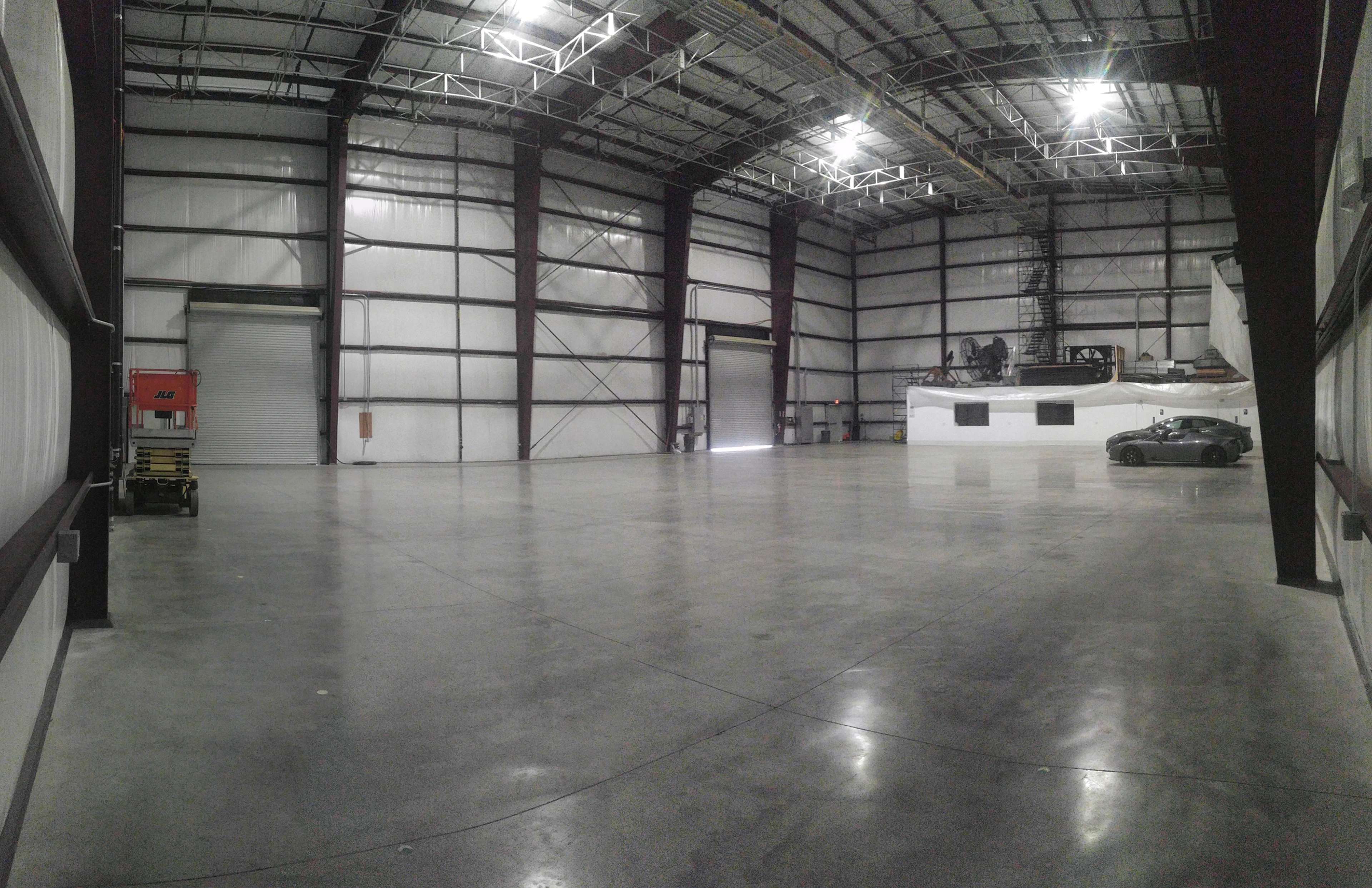 The image shows a large, empty industrial warehouse with high ceilings and a smooth concrete floor.