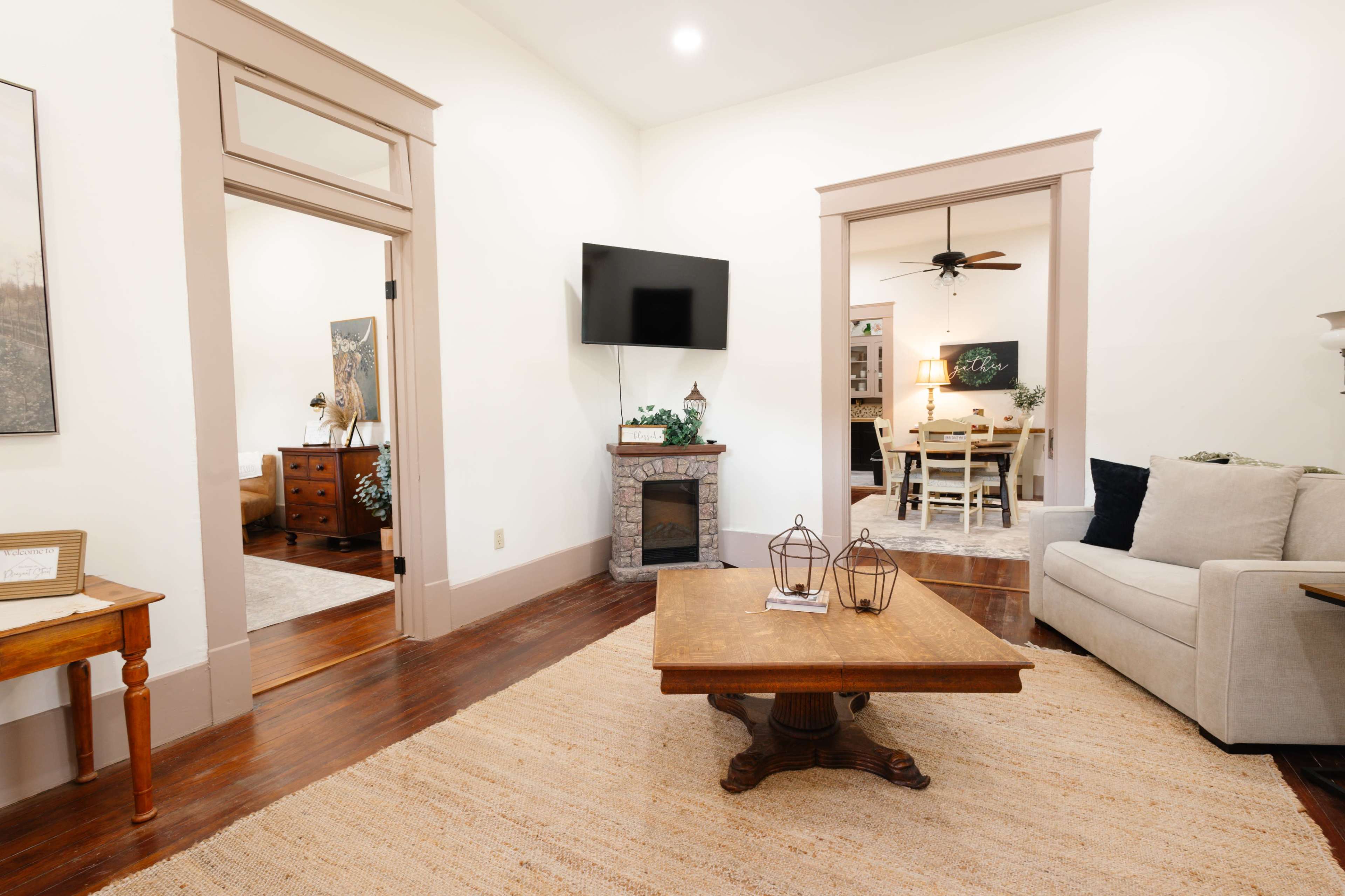 The image shows a living room featuring a beige sofa, a wooden coffee table, and doorways leading to other rooms, with a television mounted on the wall and a decorative fireplace.
