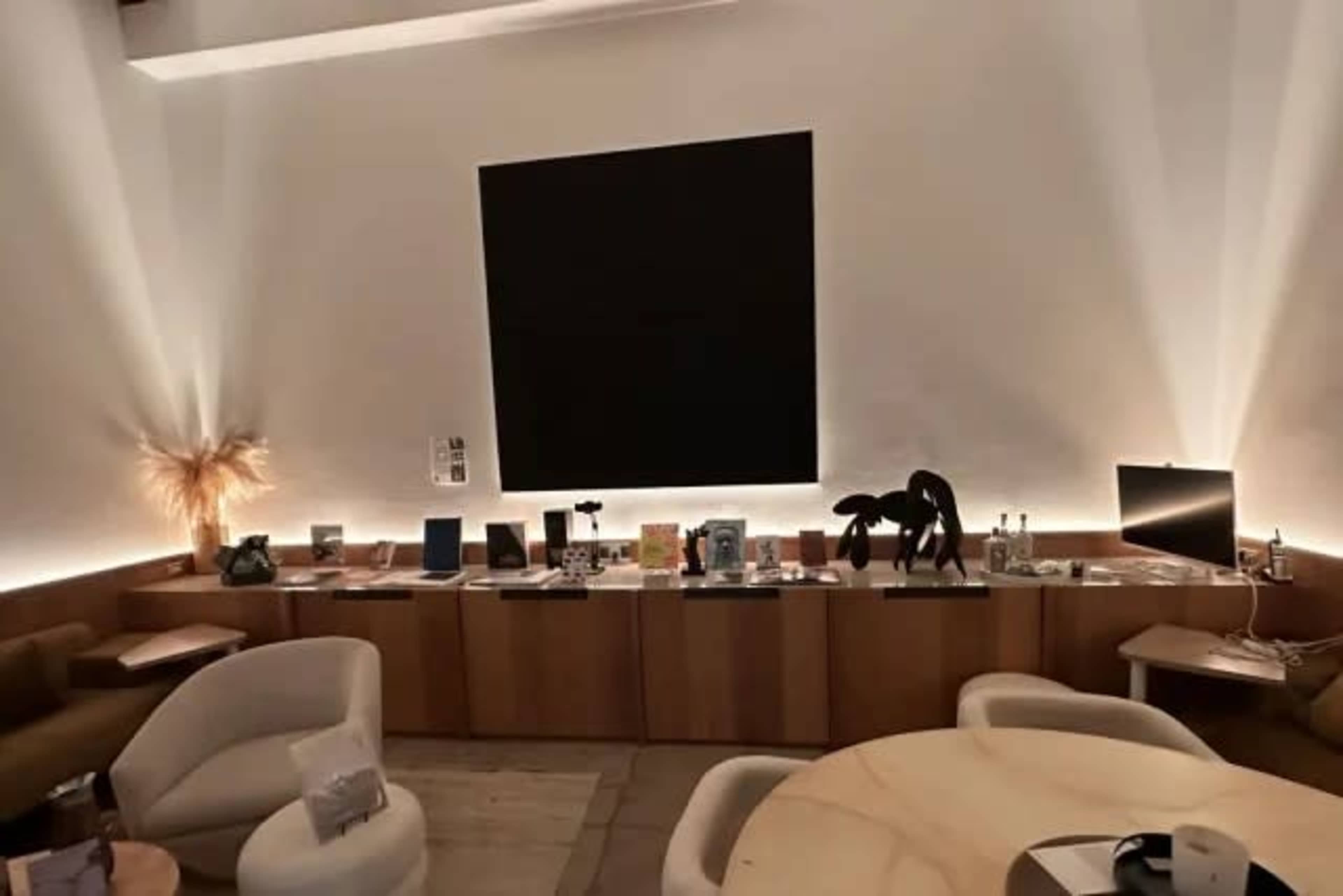 The image shows a dimly lit interior featuring a long wooden shelf with various items and books displayed, a large black wall art piece, and two seating areas.