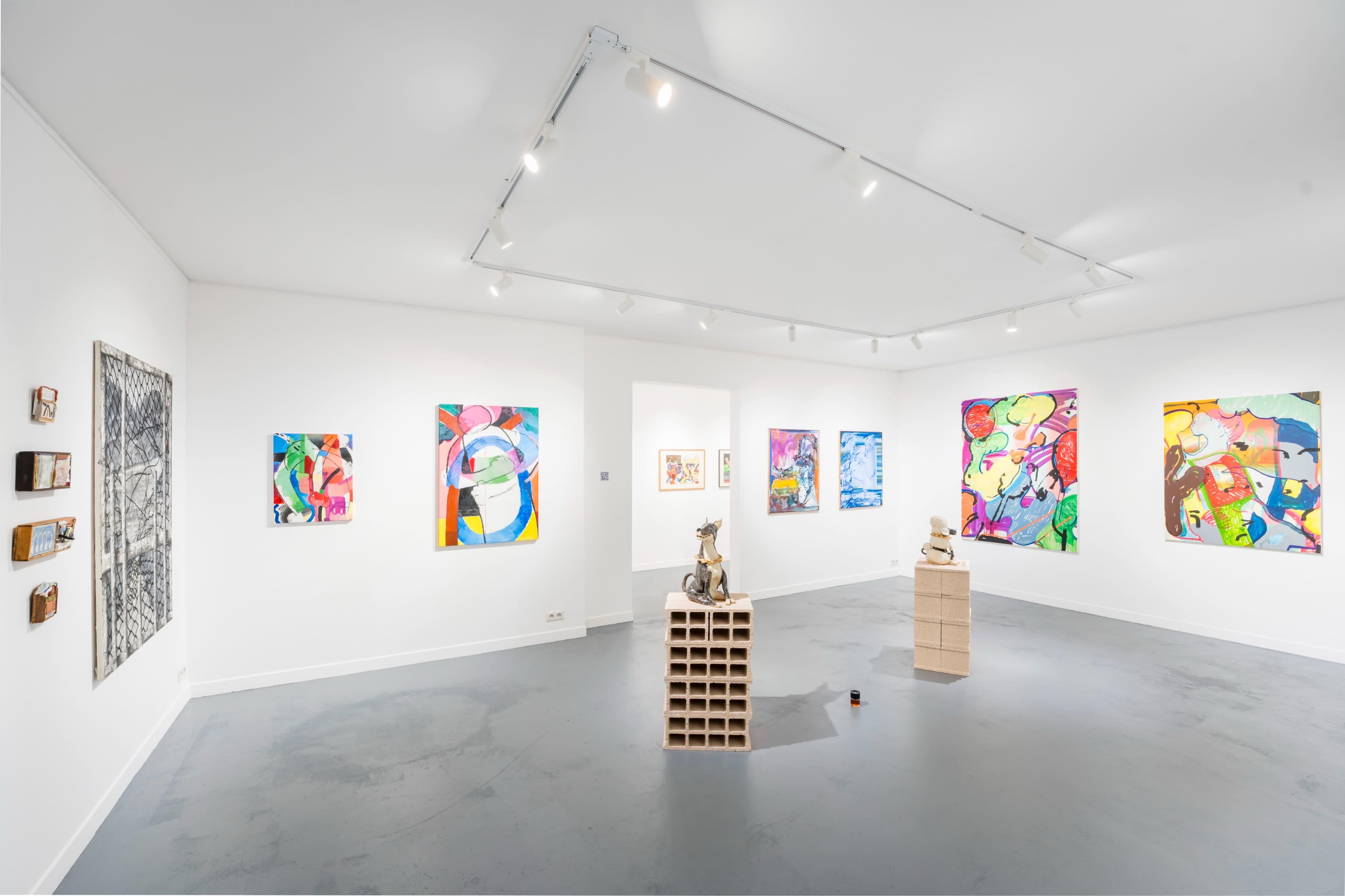 A contemporary art gallery features colorful abstract paintings and sculptures displayed on white walls and a gray floor.