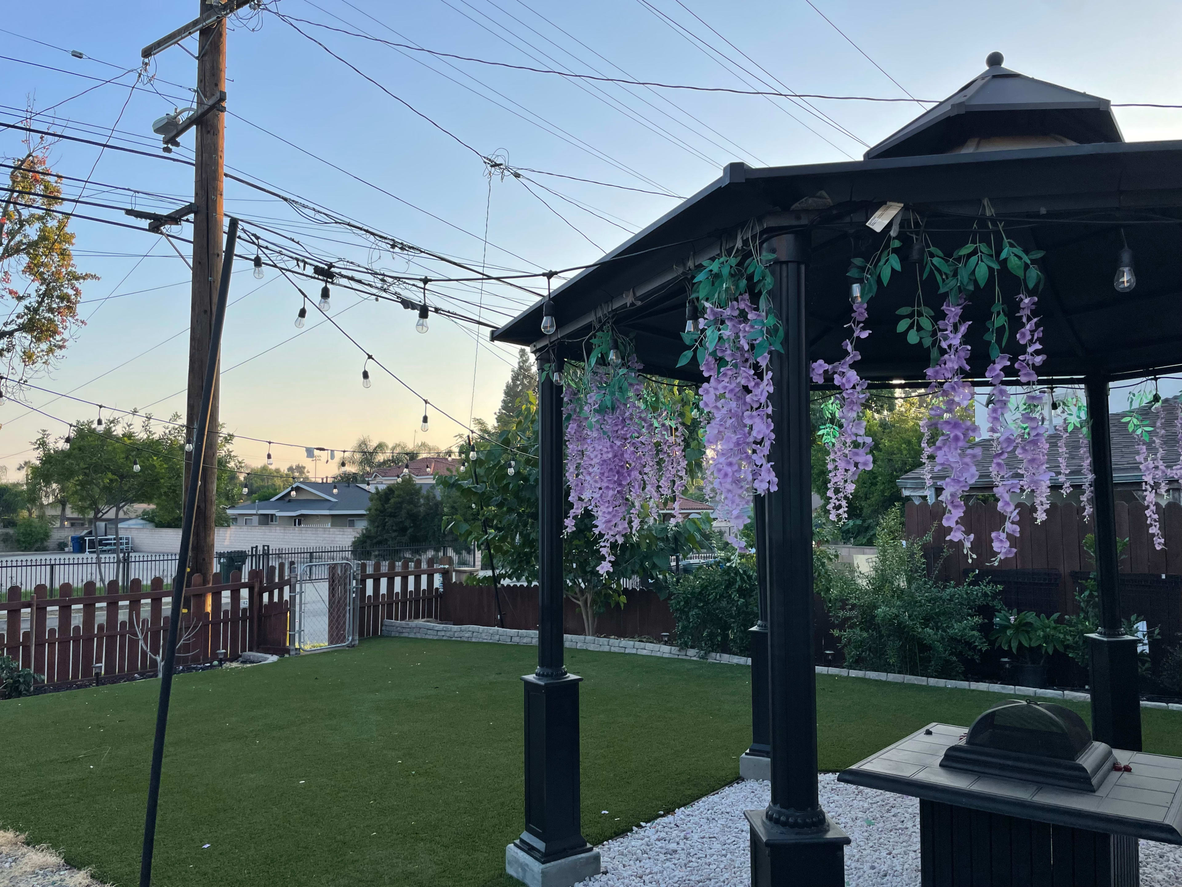 A gazebo with hanging purple flowers is situated in a backyard with artificial grass, surrounded by power lines and a fence.