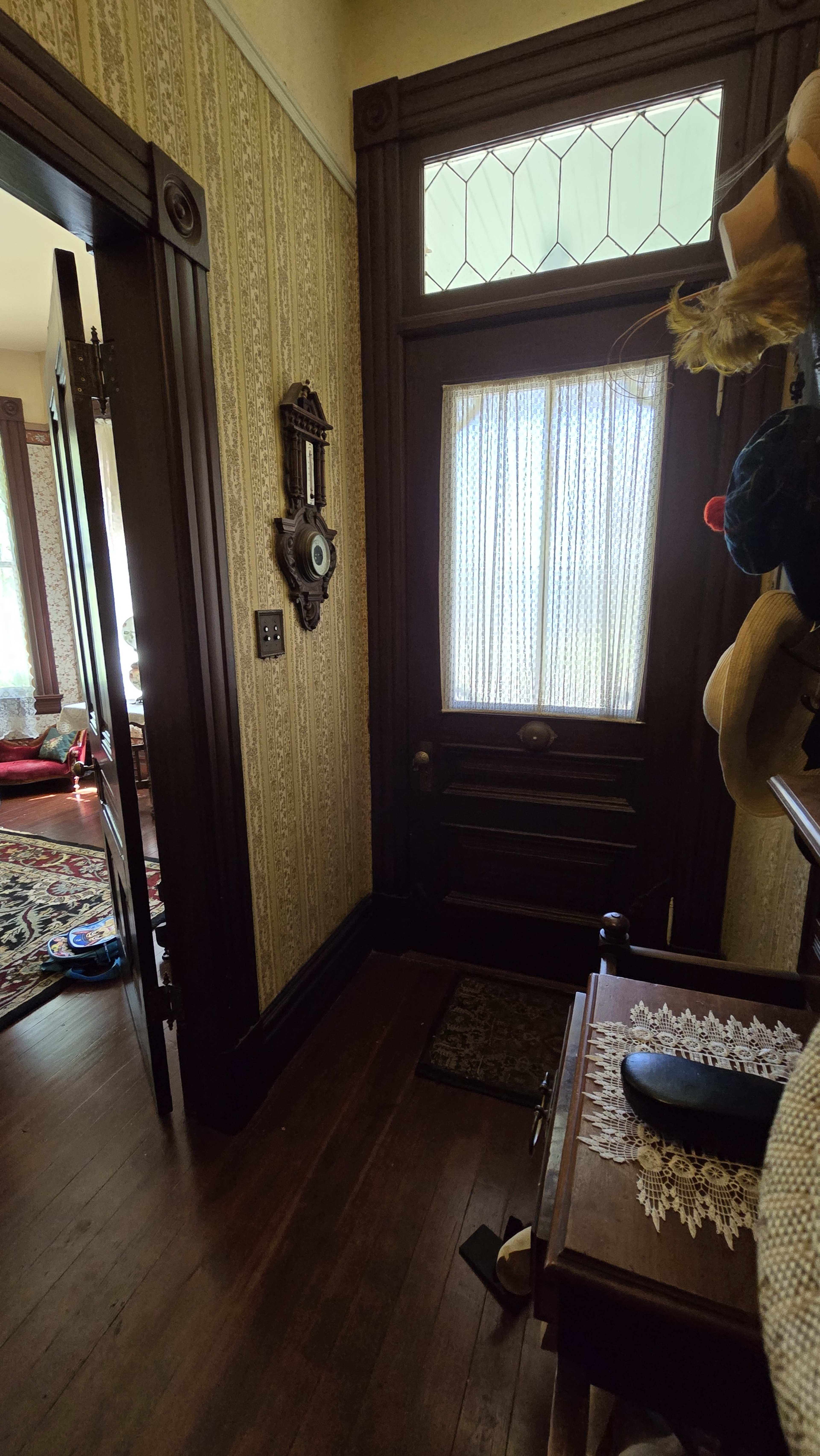A narrow hallway leading to a door with a lace curtain, flanked by ornate wooden trim and a small table decorated with a doily.