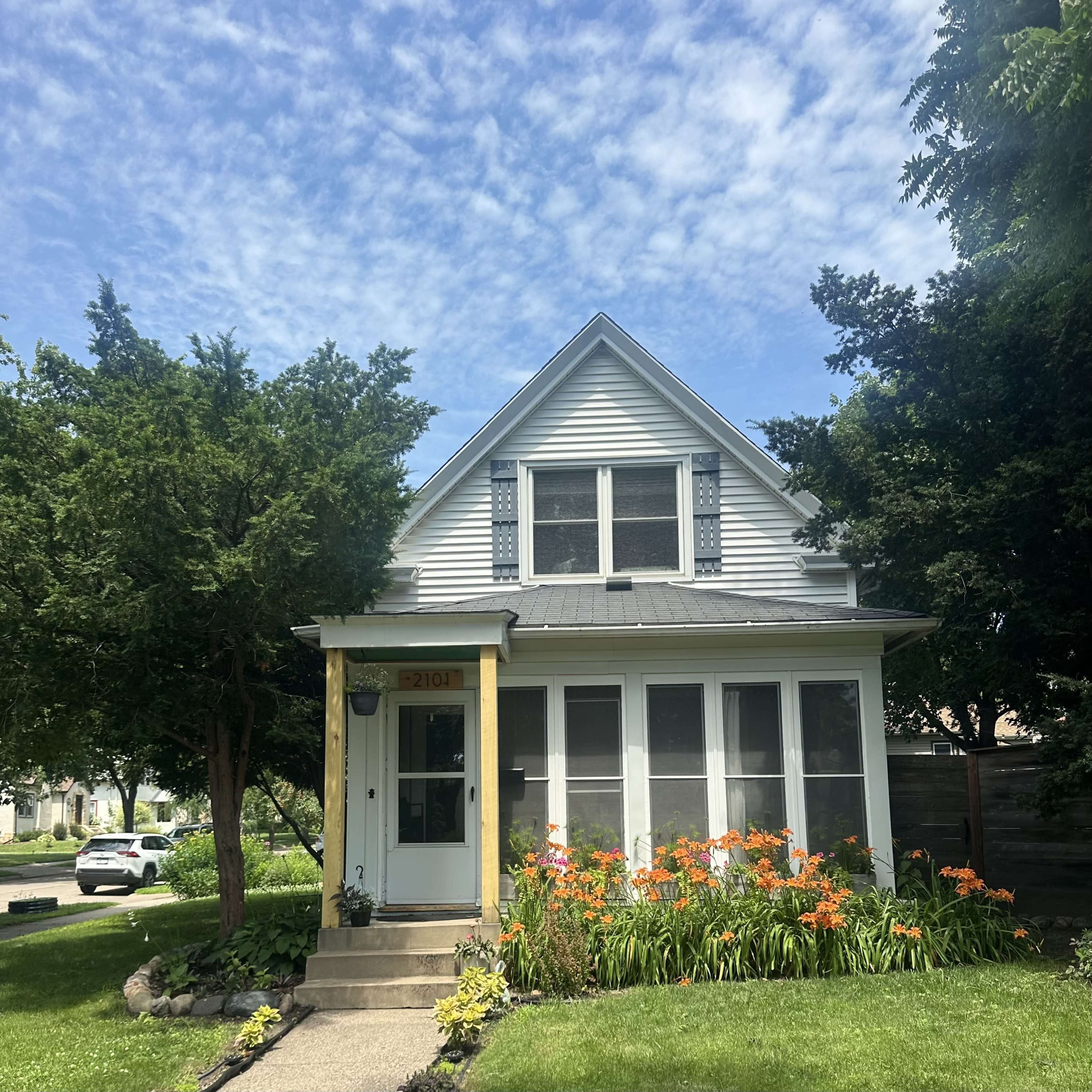 A two-story, light-colored house with a screened porch and vibrant orange flowers in the front yard is set against a blue sky with scattered clouds.
