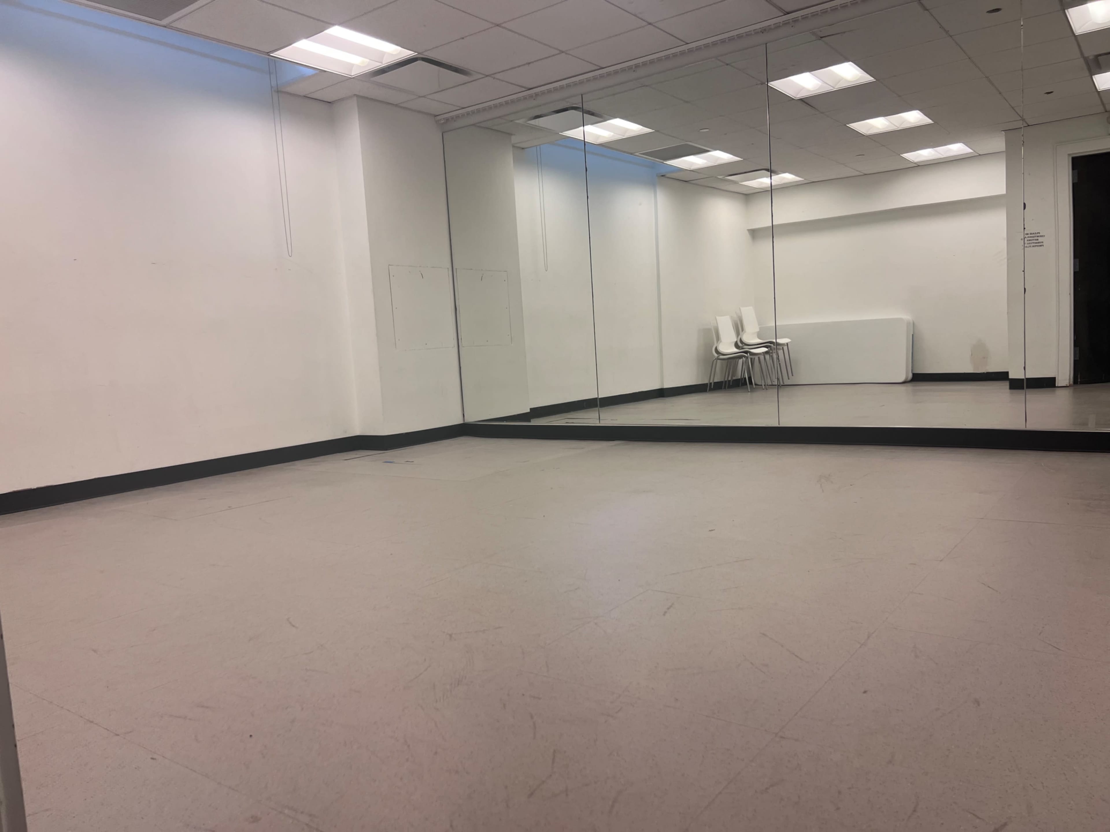 The image shows a sparsely furnished room with a mirrored wall and a few white chairs in the corner.