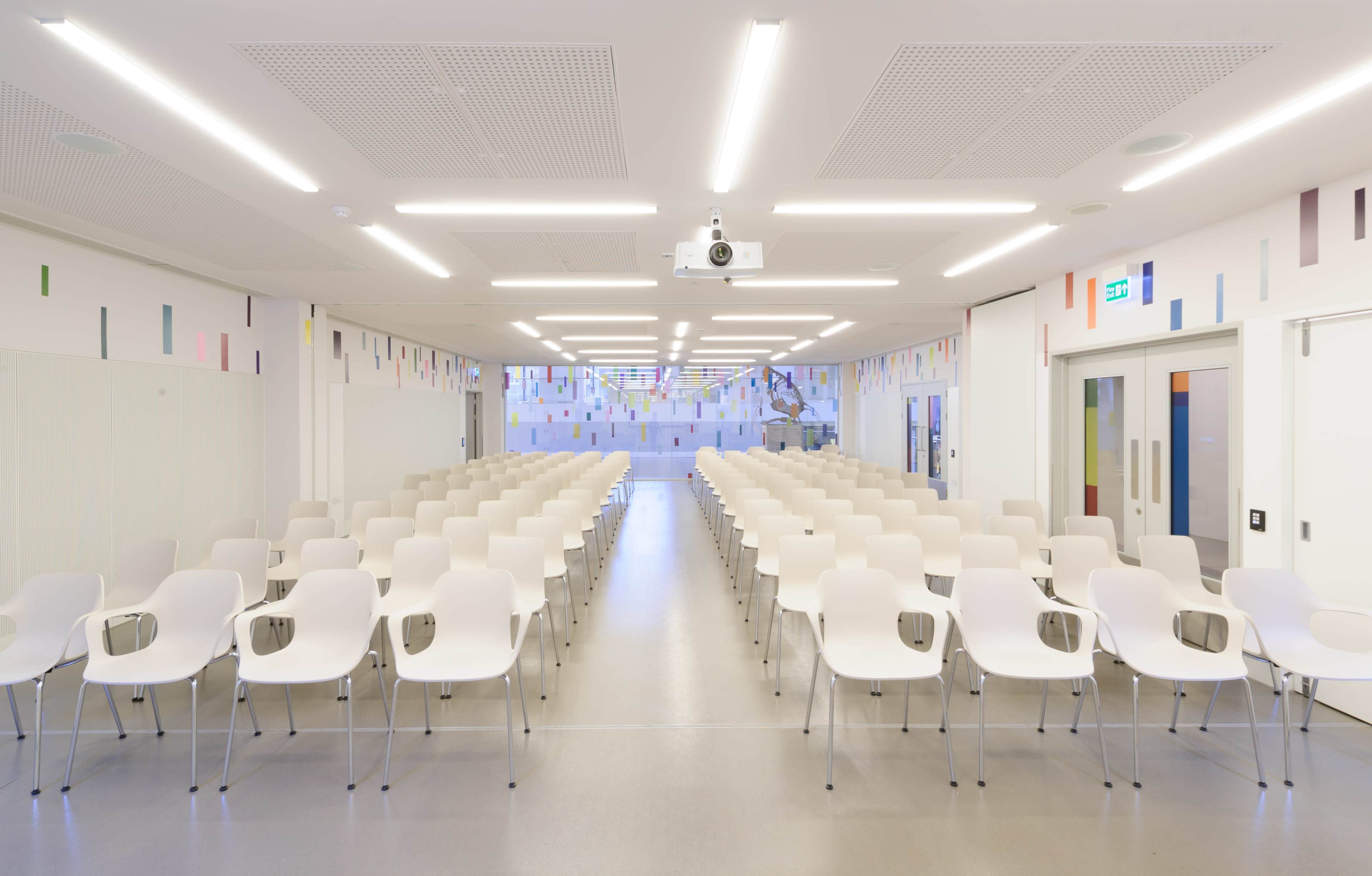 The image shows a modern conference room set up with rows of white plastic chairs facing a projection screen, with colorful decor on the walls.