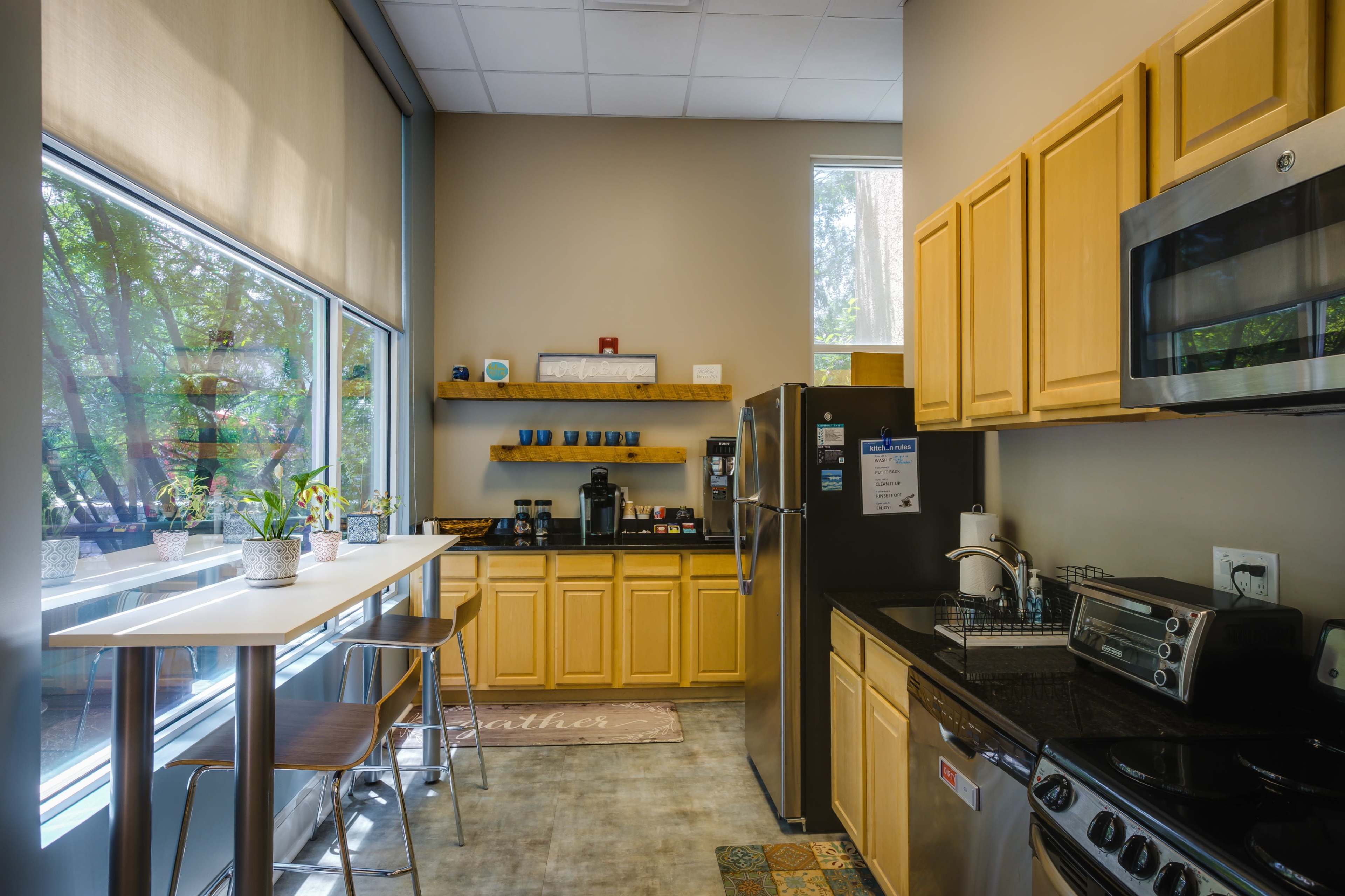 The image shows a modern kitchen with wooden cabinets, a stainless steel refrigerator, and a counter with high chairs beside large windows.
