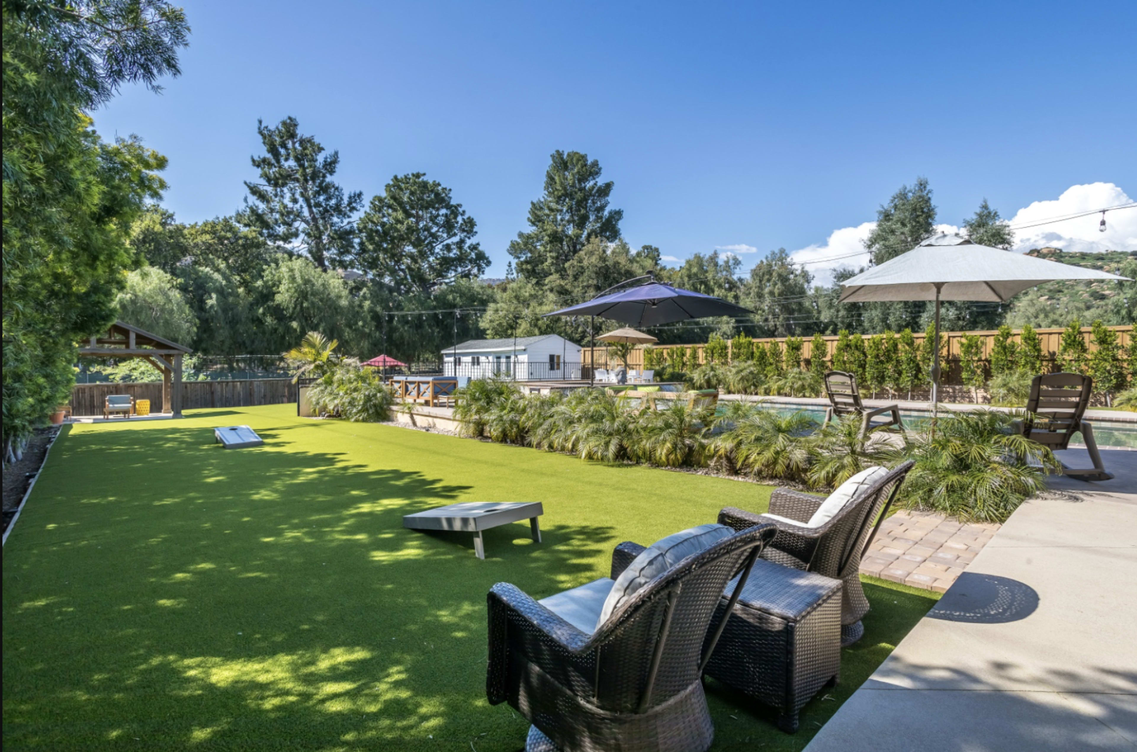 The image shows a landscaped outdoor area featuring artificial grass, lounge chairs, an umbrella, and a swimming pool surrounded by trees and a wooden fence.