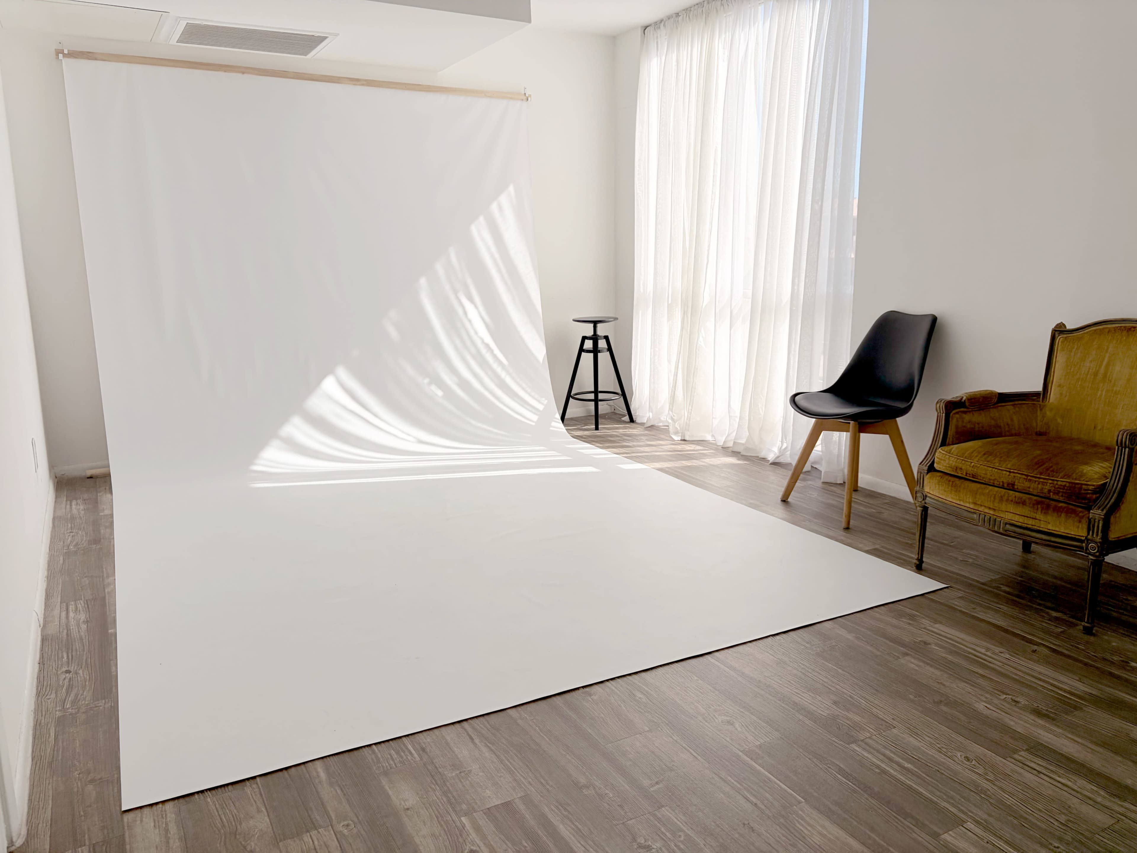 A spacious, sunlit room features a large white backdrop, a wooden floor, and two chairs, one black and one upholstered in yellow.