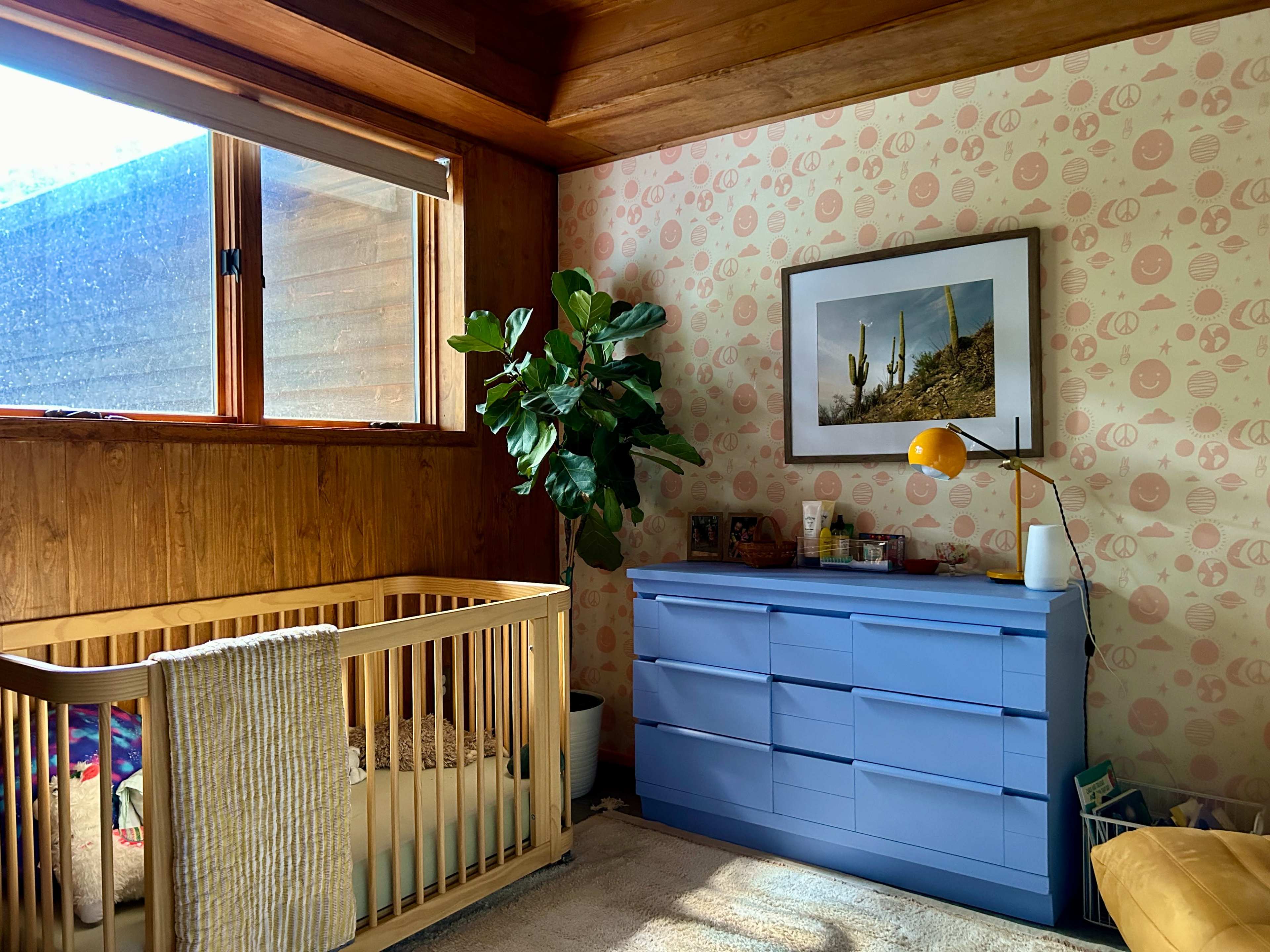 A nursery features a wooden crib, a blue dresser, a potted plant, and a framed photograph on the wall.