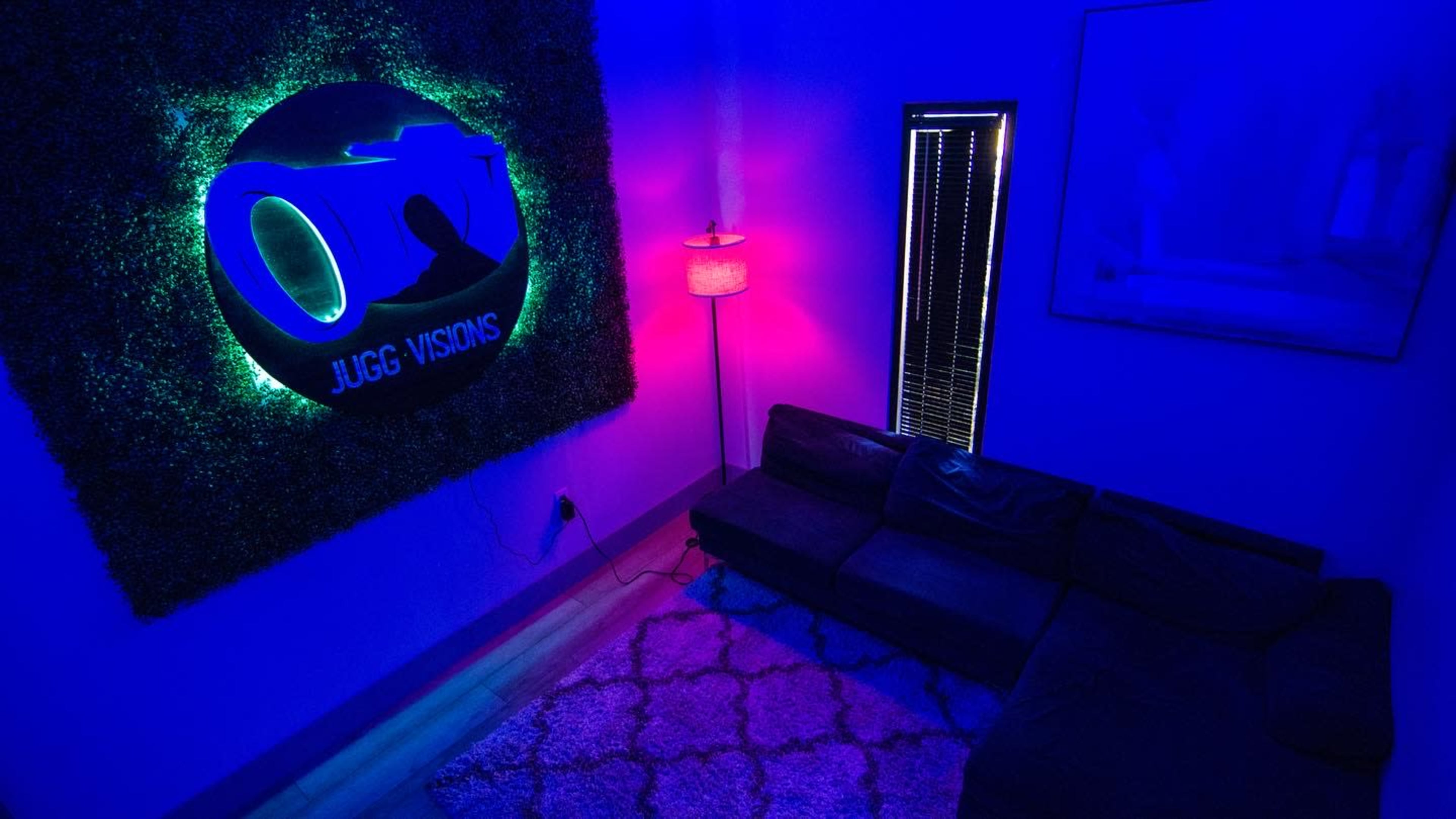A dimly lit room with blue and purple lighting, featuring a neon sign that reads "JUGG VISIONS" on a textured wall, a lamp beside a dark sofa, and a window with blinds.