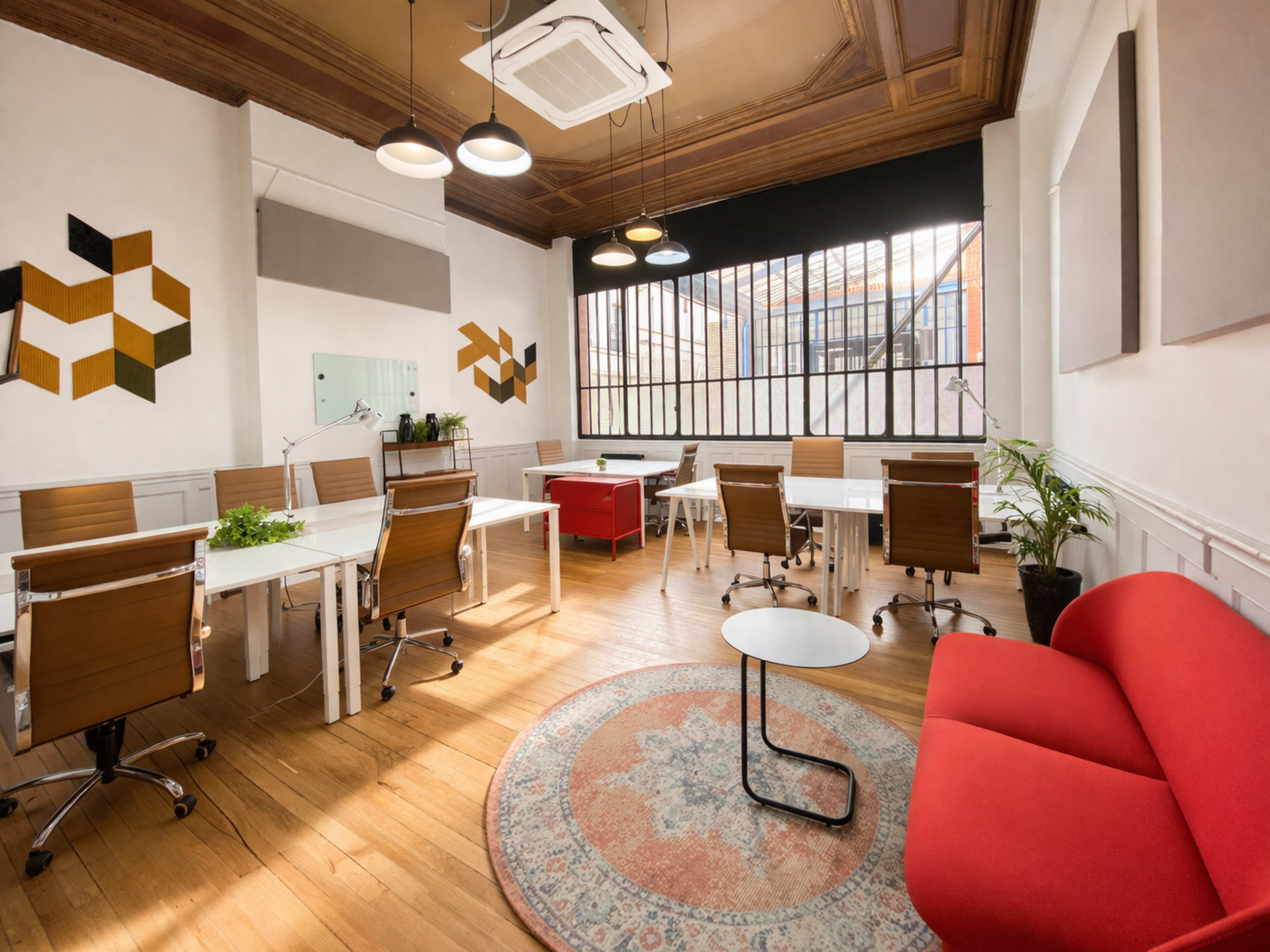 The image shows a modern workspace with several desks, ergonomic chairs, a red seating area, and large windows allowing natural light to fill the room.