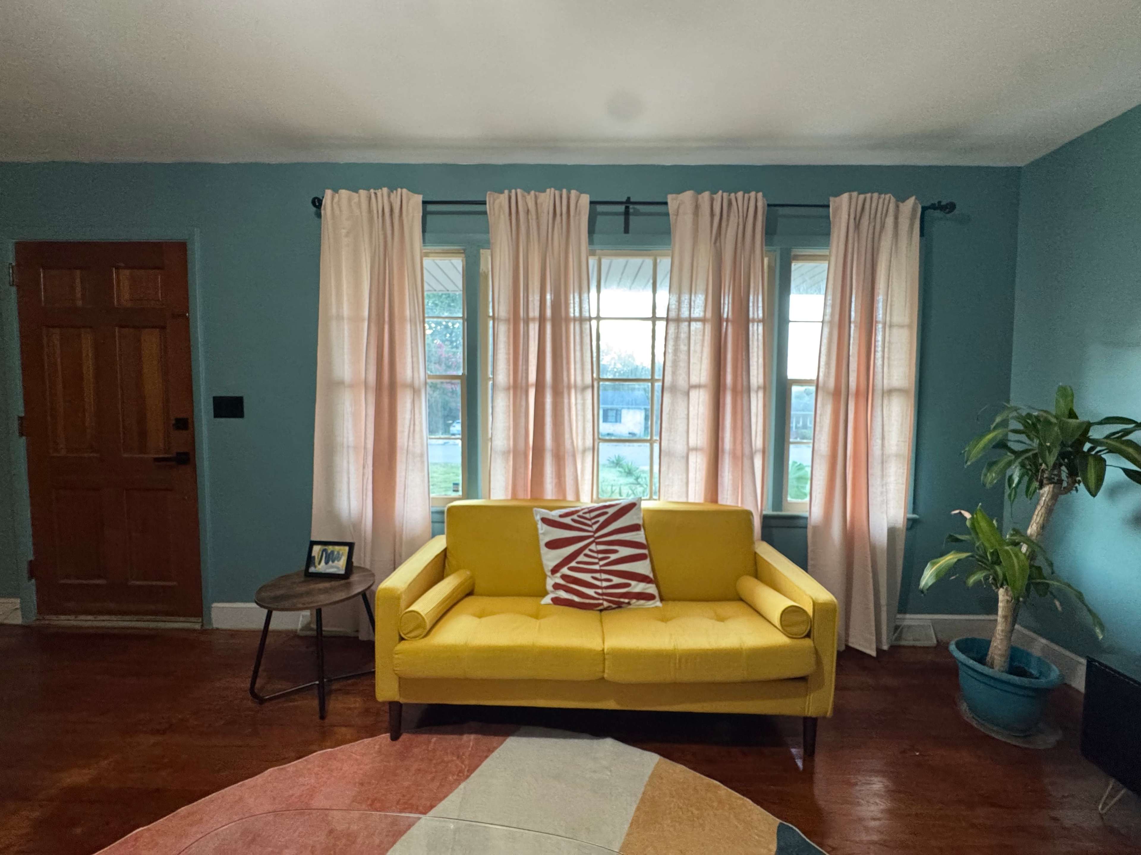 A yellow couch sits in a brightly colored room with teal walls and large windows covered by light pink curtains.