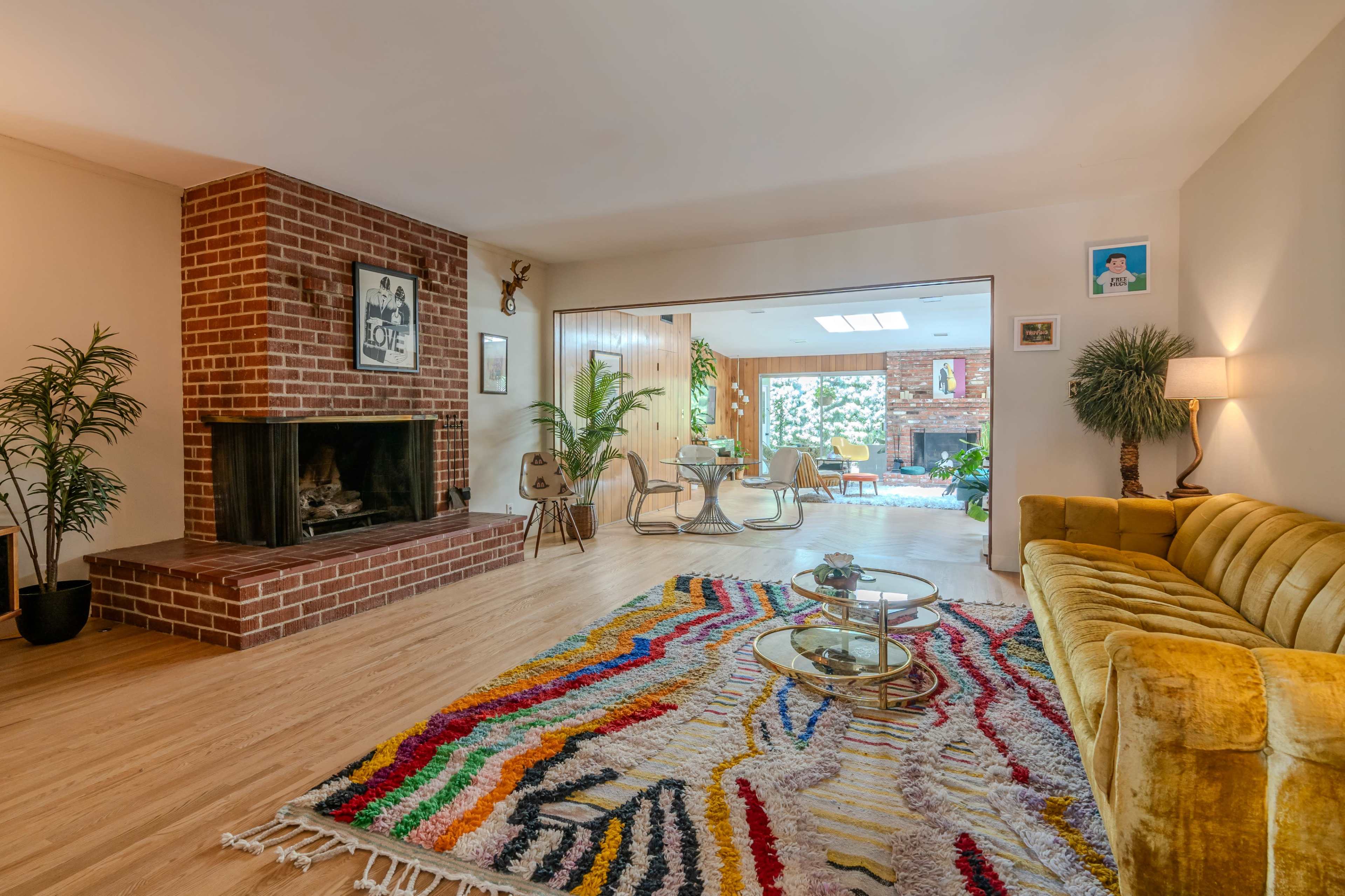 The image shows a bright, open-concept living area with a brick fireplace, a yellow couch, and a colorful area rug, leading to a dining space with large windows.