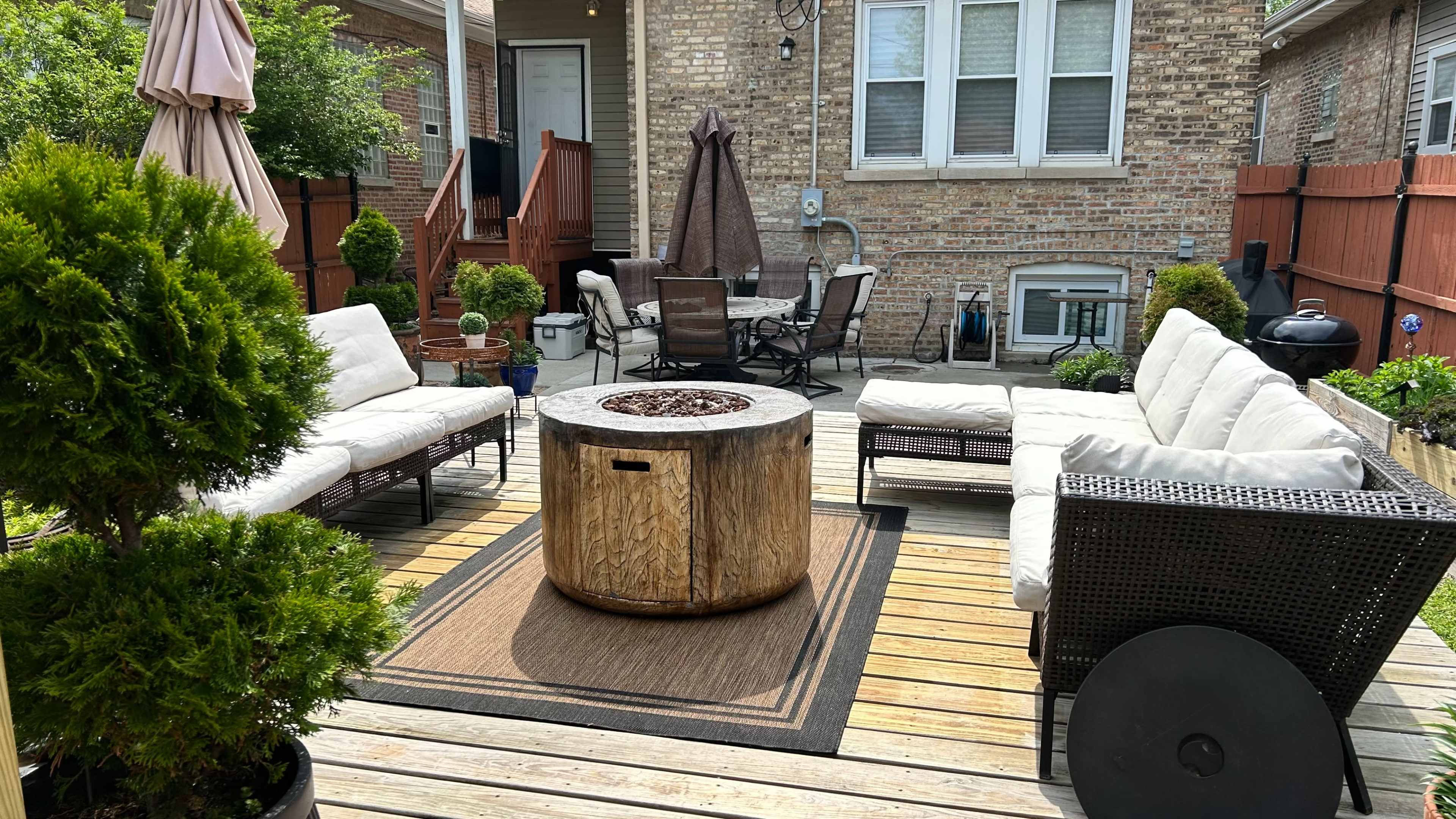 The image shows a backyard deck area with a circular wooden fire pit at the center, surrounded by seating areas made of white cushions and wicker furniture.