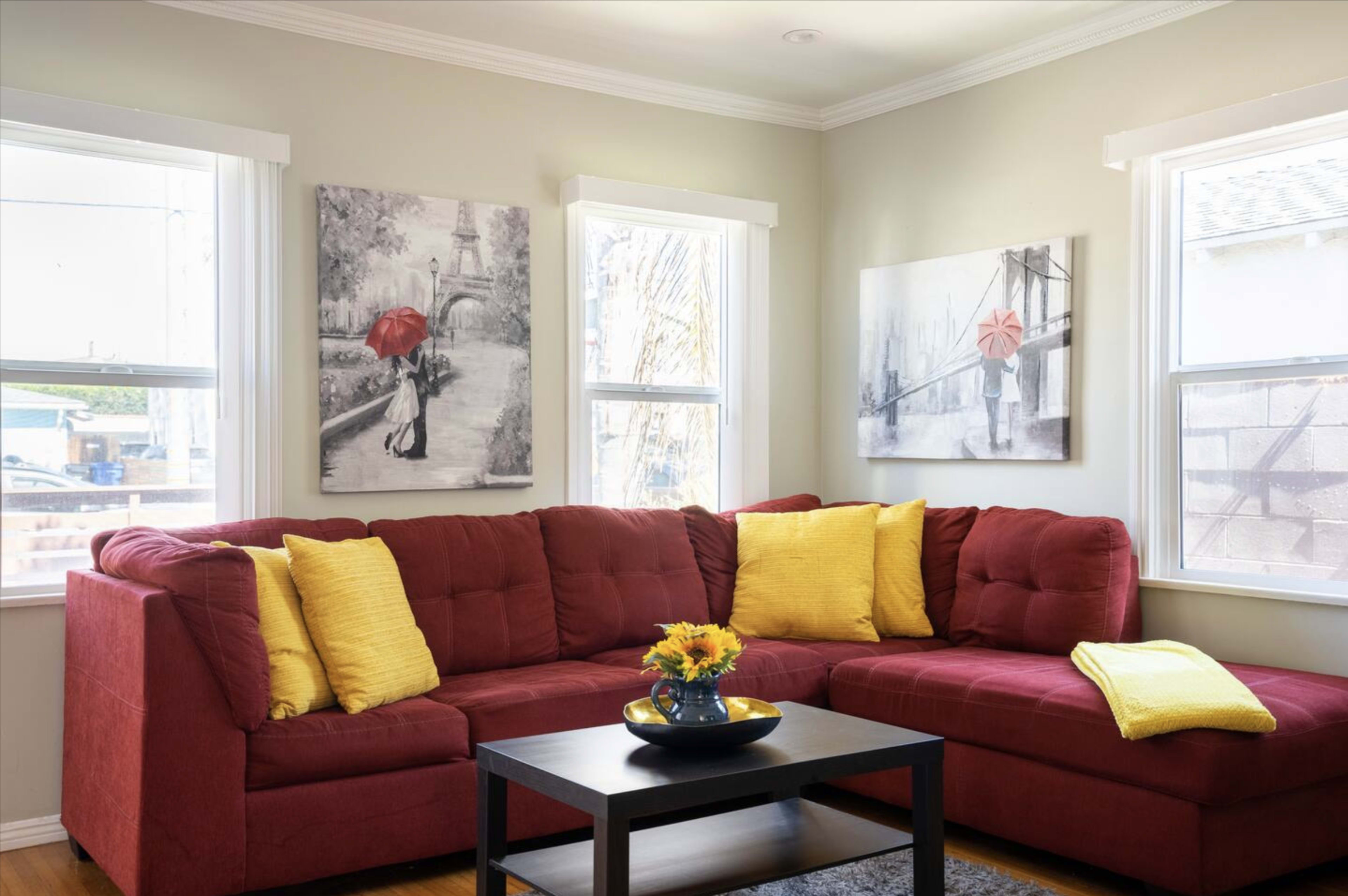 A red sectional sofa with yellow pillows is positioned next to a black coffee table featuring a vase of sunflowers in a well-lit living room adorned with framed artwork on the walls.