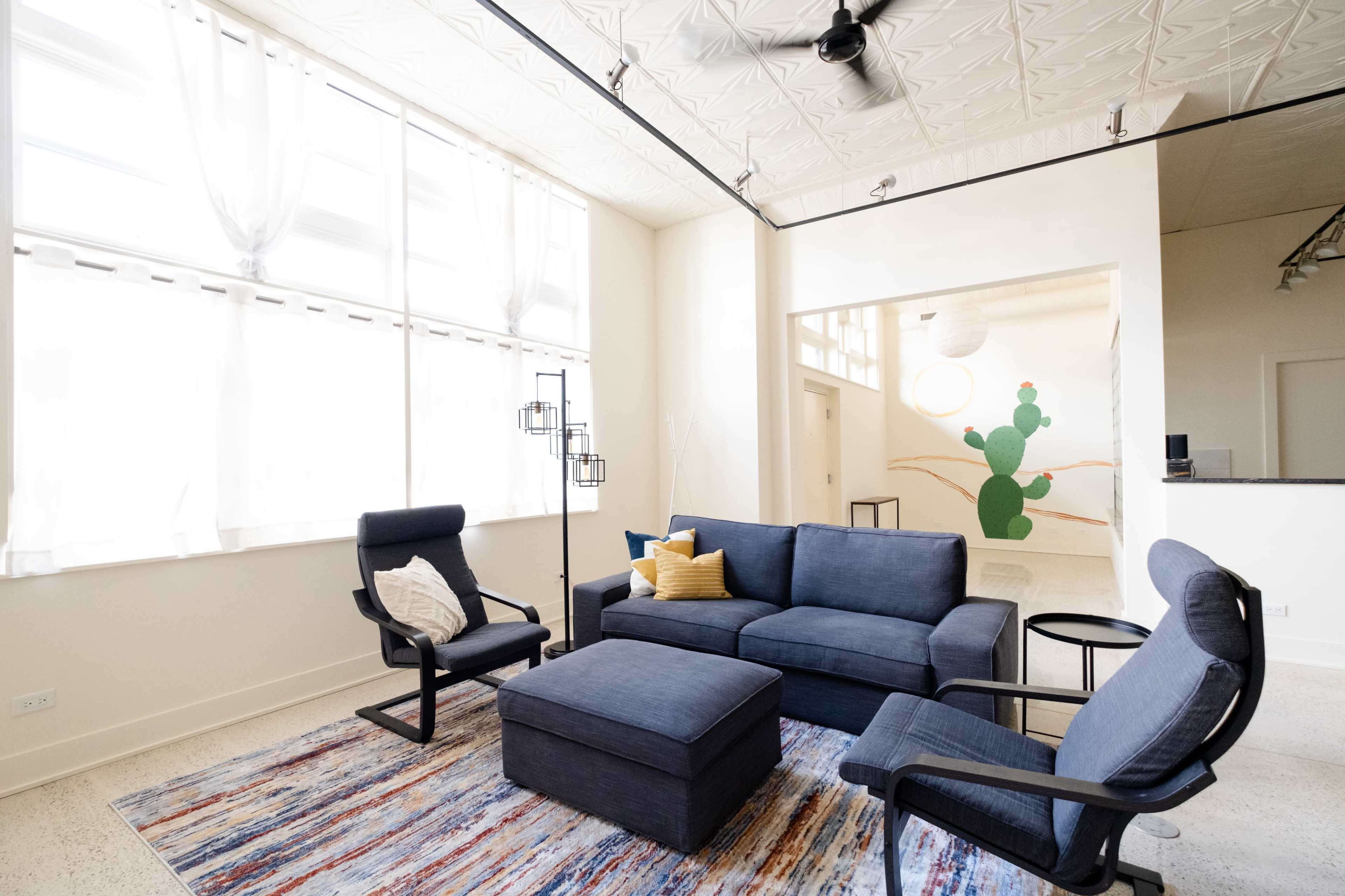 A modern living room features a navy blue sofa and chairs, a patterned rug, large windows with light curtains, and a green cactus mural in the background.