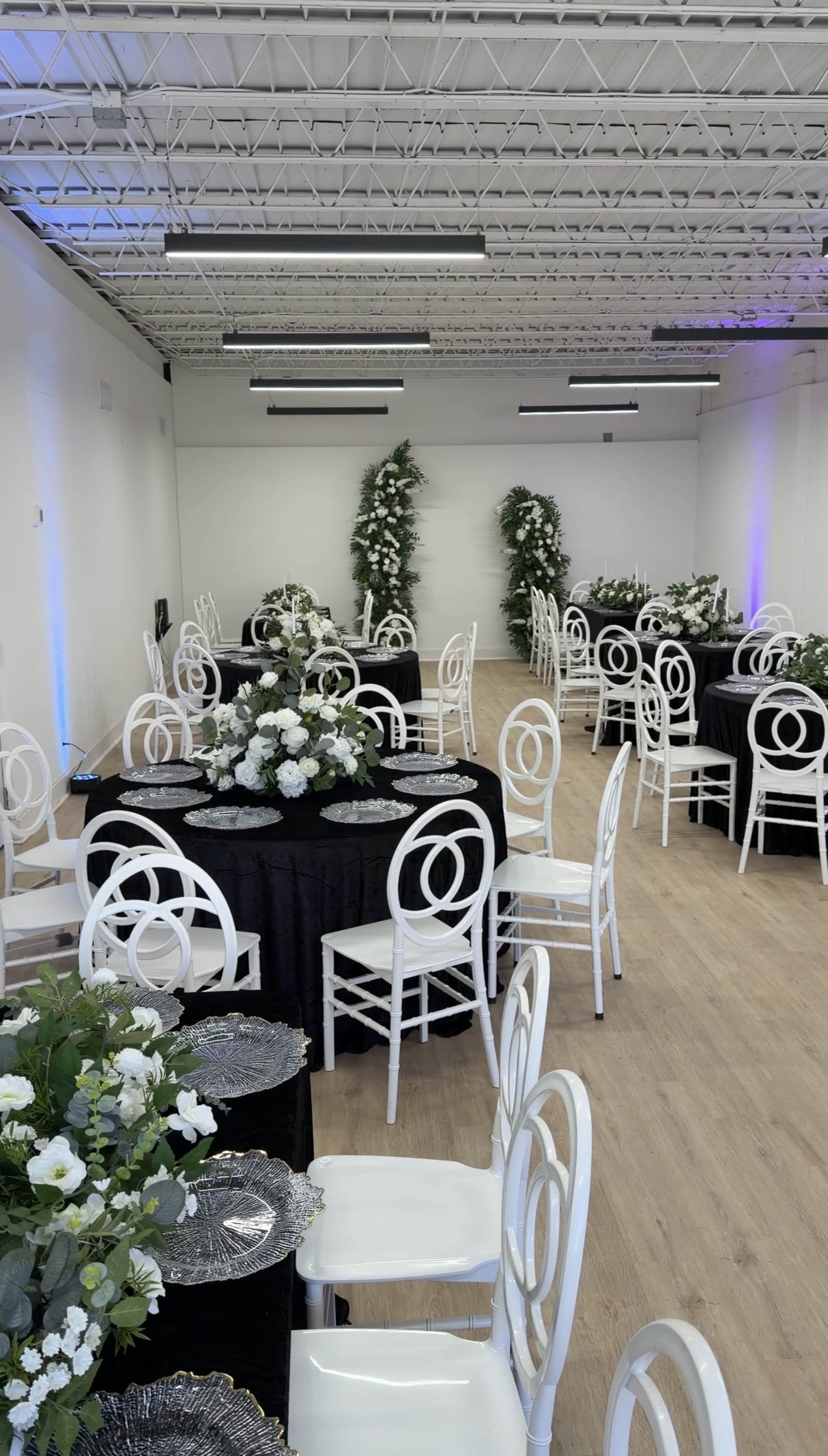 The image shows a modern event space with neatly arranged tables covered in black linens, white chairs, and floral centerpieces.