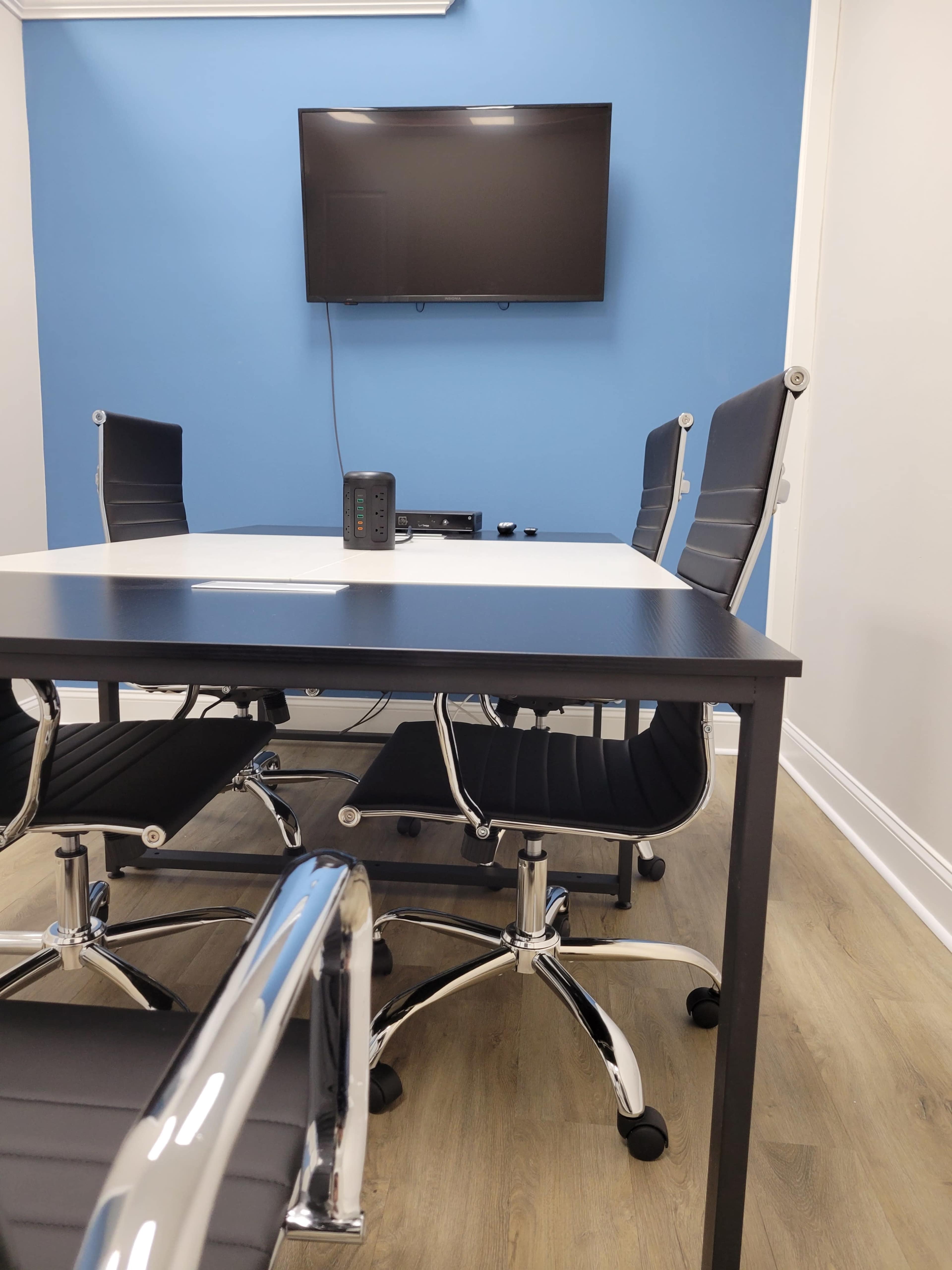 A modern conference room with a rectangular table, four ergonomic chairs, and a wall-mounted TV on a blue wall.