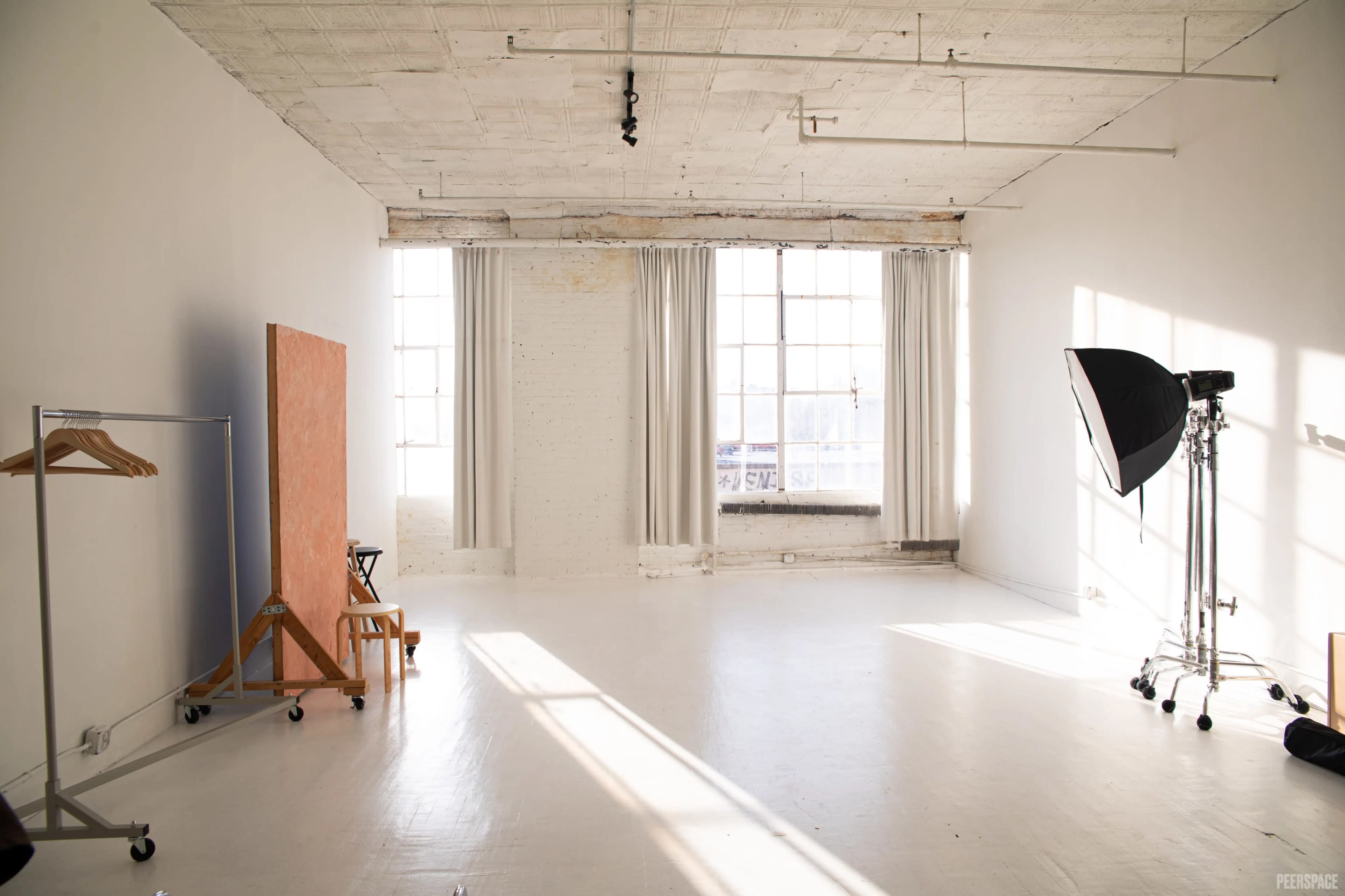 The image shows a bright, spacious room with large windows featuring white walls and an empty floor, along with a small rolling clothing rack and a backdrop stand.