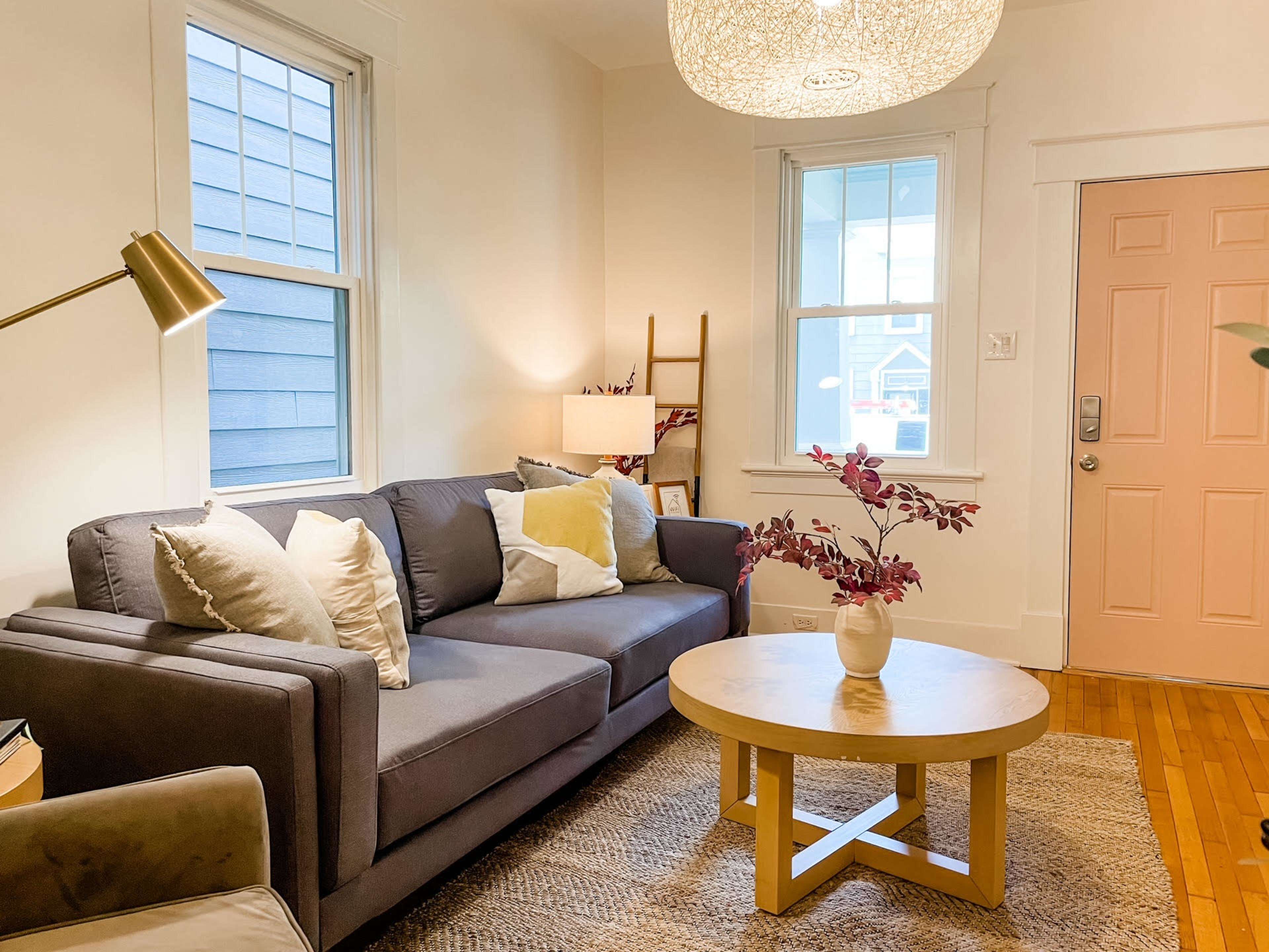 The living room features a gray sofa with decorative pillows, a round coffee table, and a large pendant light, set against light walls and wooden flooring.